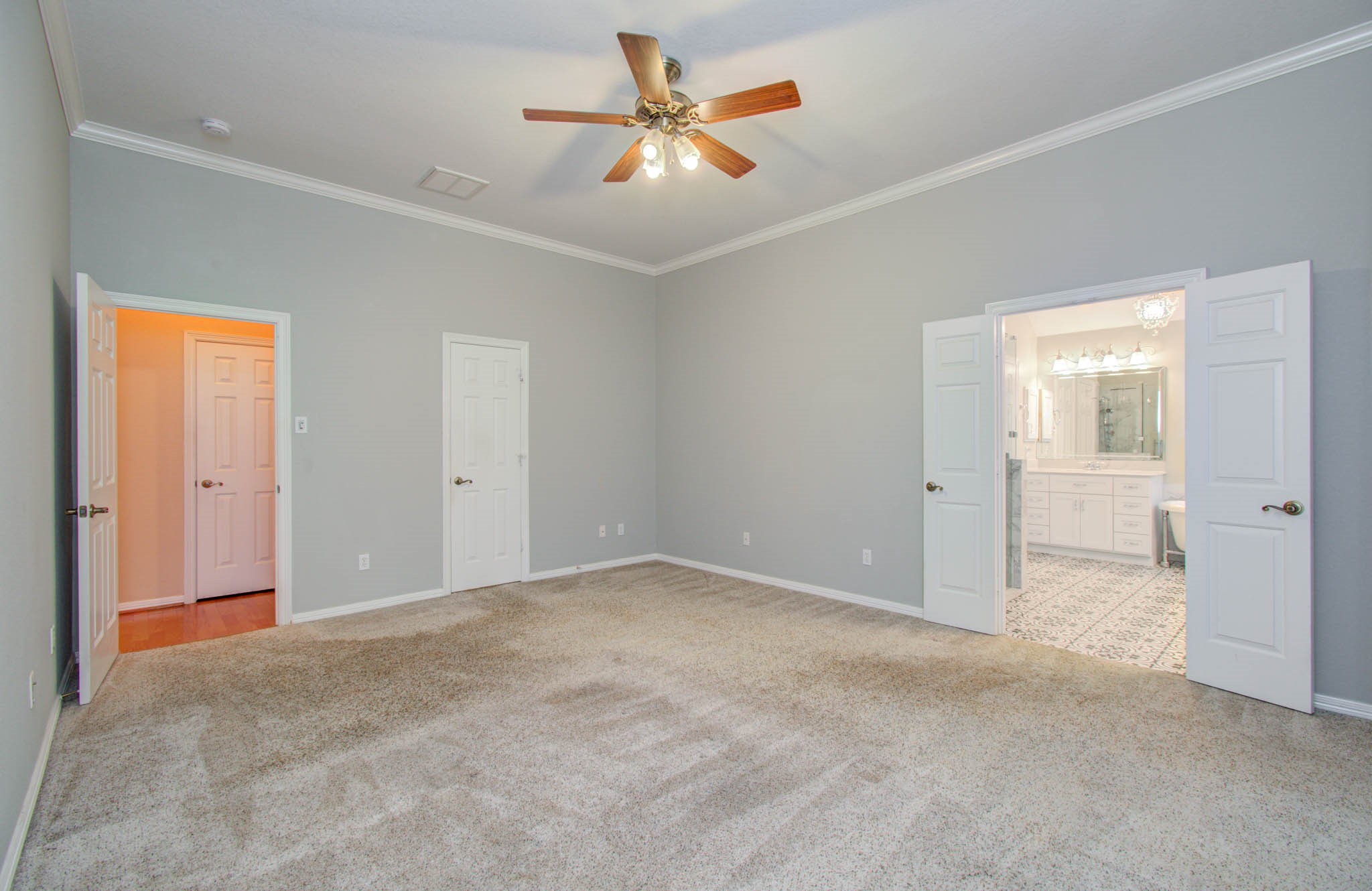 Spacious bedroom with soft carpeting, elegant crown molding, and ceiling fan, leading to a luxurious en-suite bathroom through double doors.
