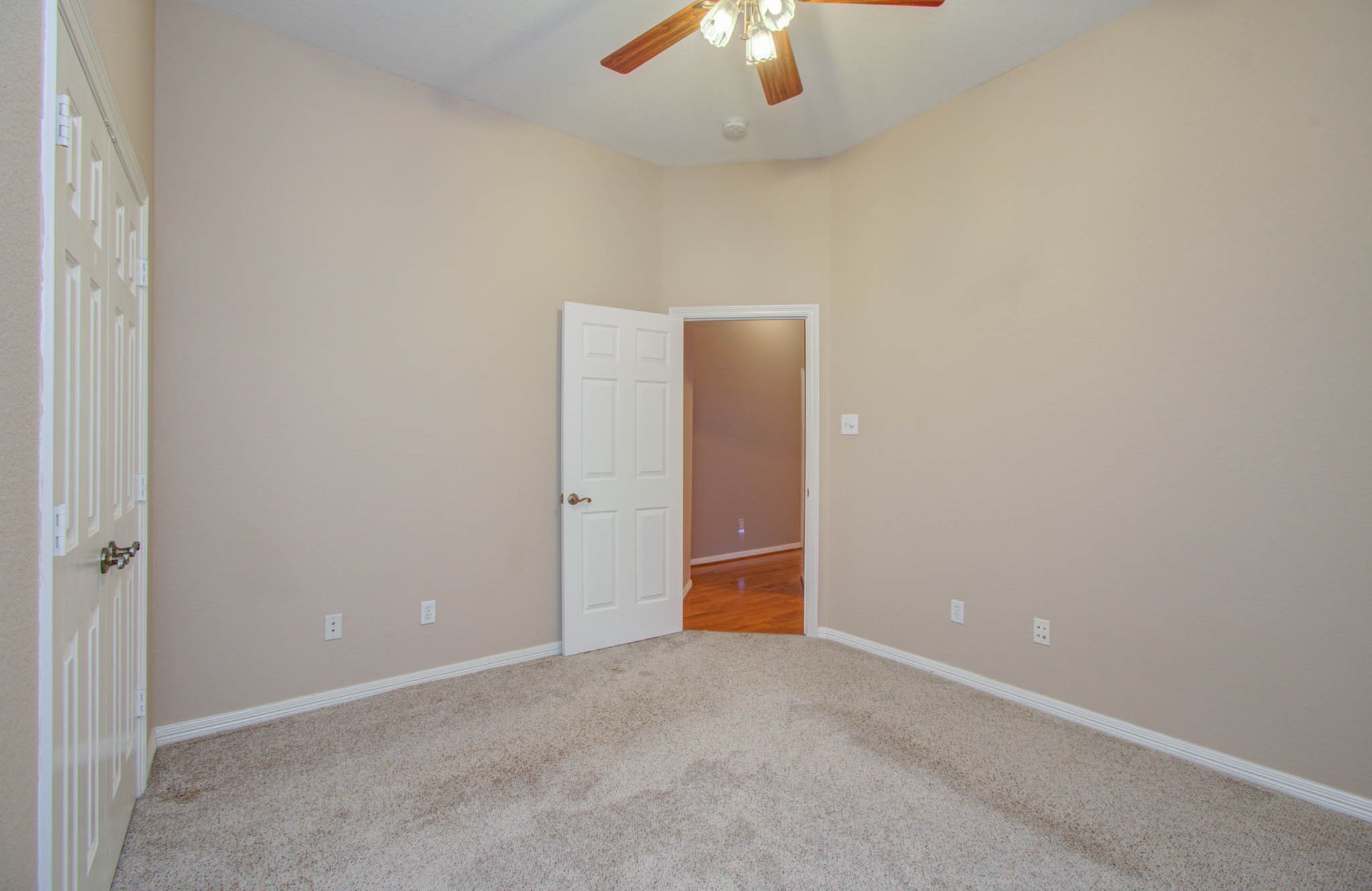 Bright bedroom with soft carpet, neutral walls, and a ceiling fan, featuring ample closet space and a doorway leading to hardwood floors.
