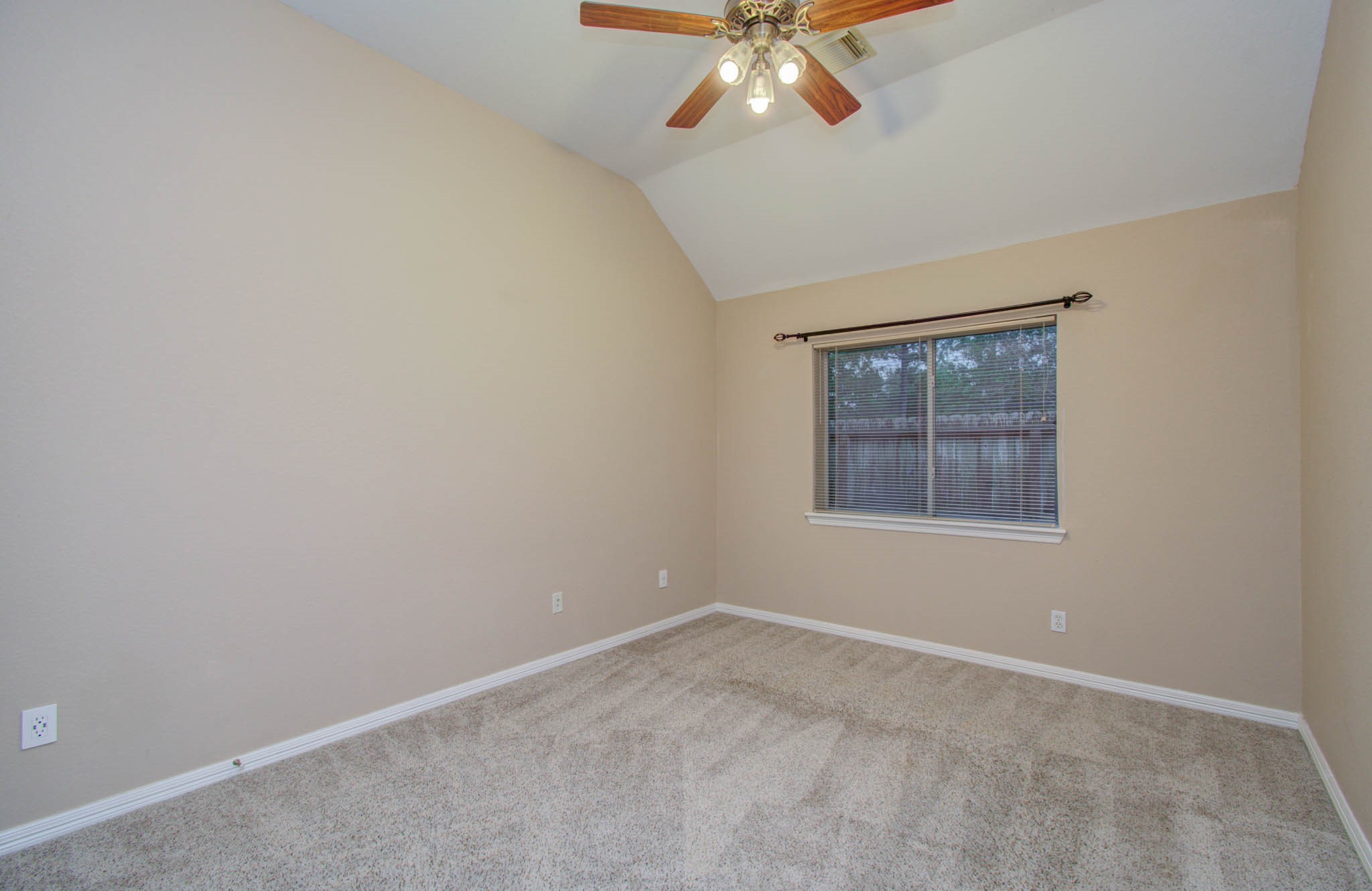Cozy bedroom with soft carpeting, neutral walls, and a vaulted ceiling featuring a stylish ceiling fan. Natural light pours in through the window, creating a warm and inviting atmosphere.