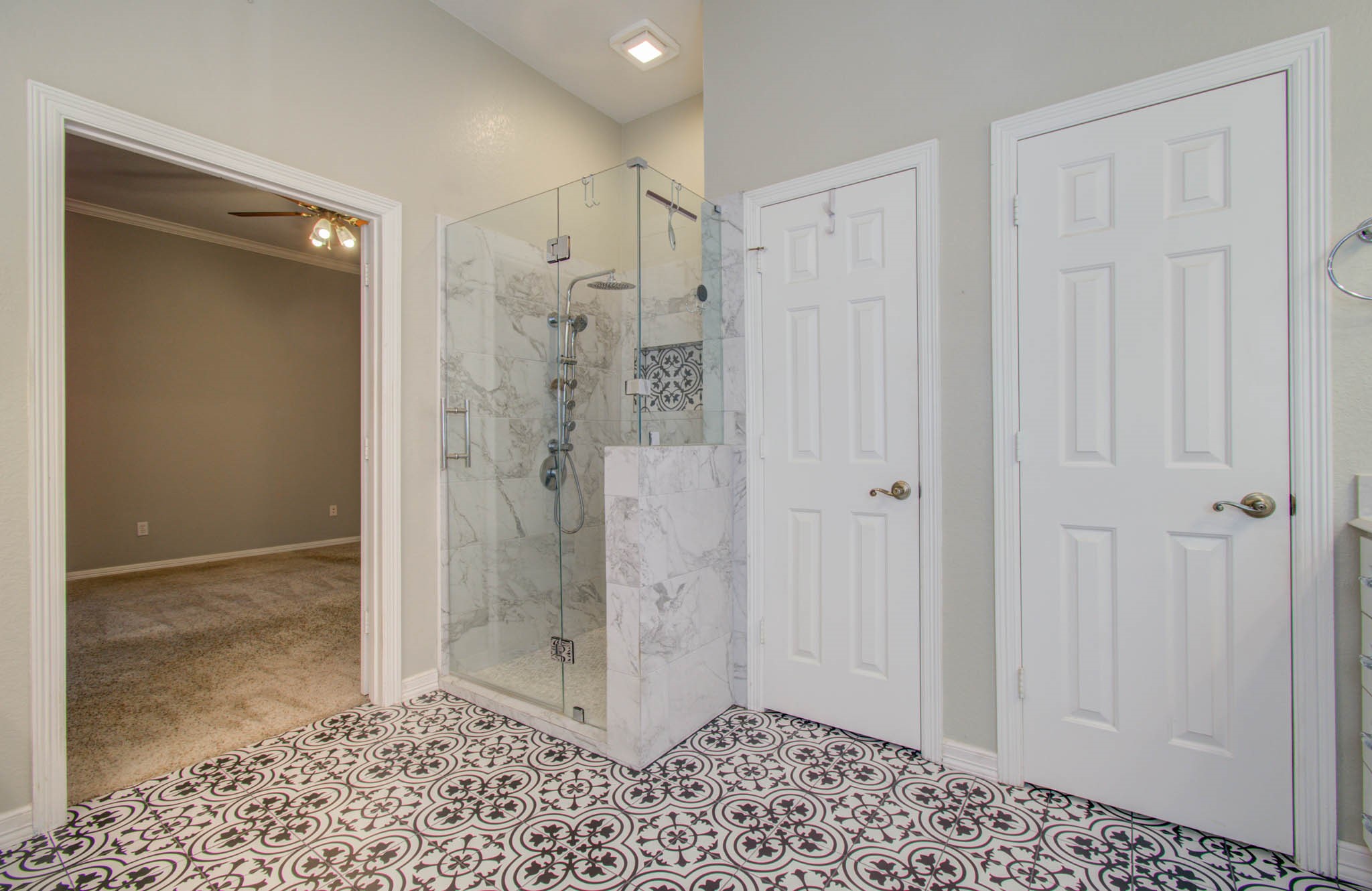 Elegant bathroom featuring a luxurious glass-enclosed marble shower and stylish patterned tile flooring, with convenient access to an adjoining carpeted room.