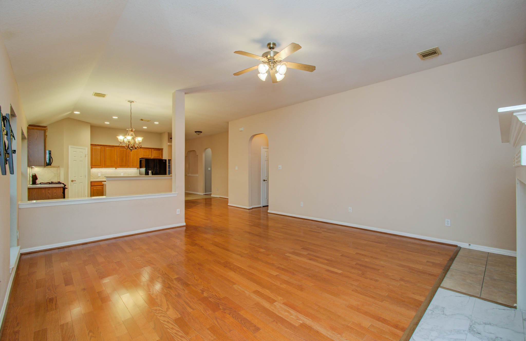 Spacious living area with warm hardwood floors, high ceilings, and a ceiling fan, seamlessly flowing into an inviting kitchen with modern lighting and ample cabinetry.