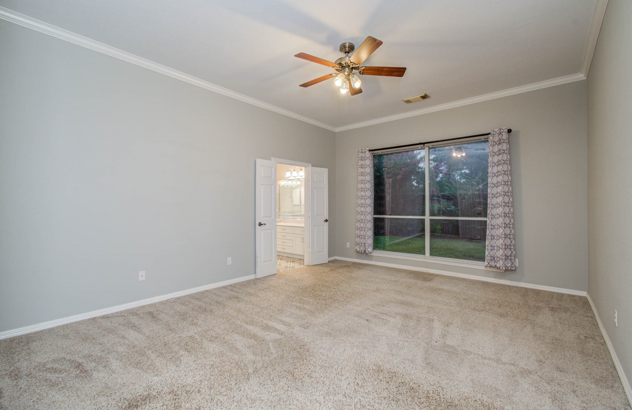 Spacious bedroom with plush carpeting, featuring a large window with elegant drapes offering plenty of natural light and a ceiling fan for comfort. An en-suite bathroom with a dual vanity is accessed through double doors.