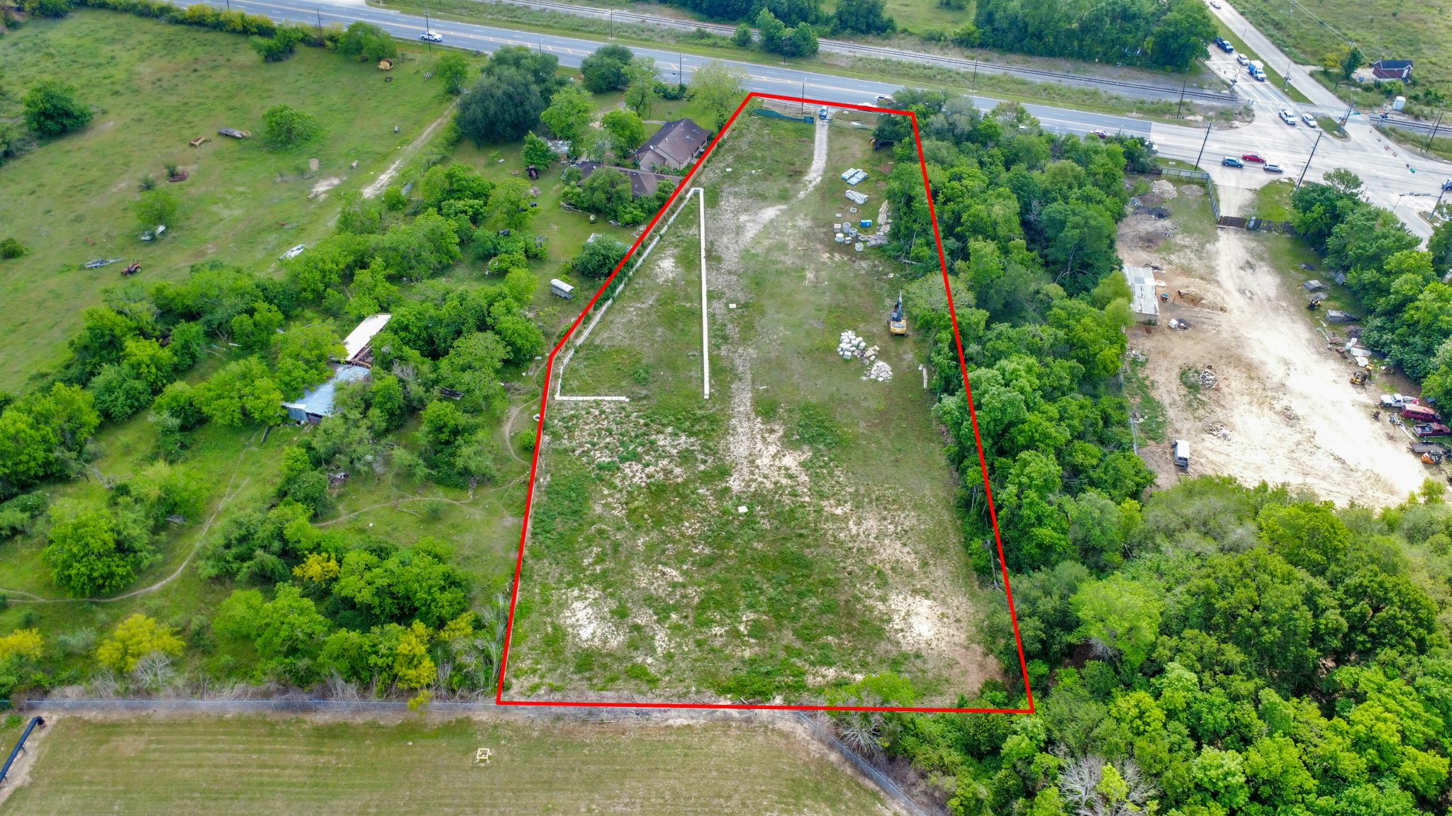 This photo shows a large, triangular plot of land outlined in red, featuring a mix of open grassy areas and scattered trees. It's adjacent to a road, offering easy access, with some nearby buildings and open spaces nearby, suggesting potential for development or agricultural use.