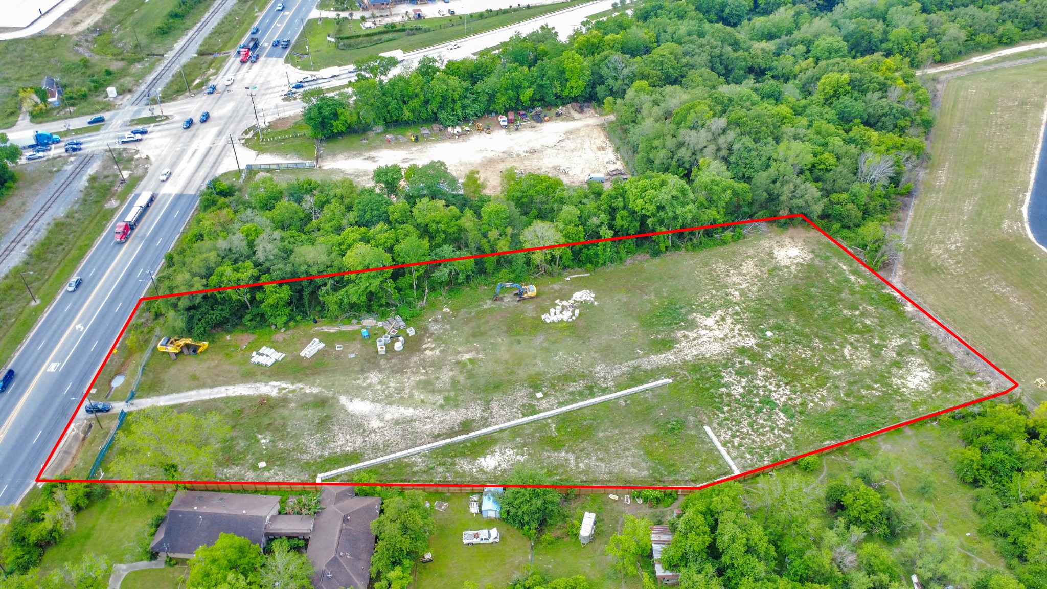 This aerial photo shows a large, triangular plot of land bordered by trees and a busy road on one side. The area is mostly open with some construction materials and machinery, suggesting potential for development. The plot is outlined in red, highlighting its boundaries. This aerial photo shows a large, triangular plot of land bordered by trees and a busy road on one side. The area is mostly open with some construction materials and machinery, suggesting potential for development. The plot is outlined in red, highlighting its boundaries.