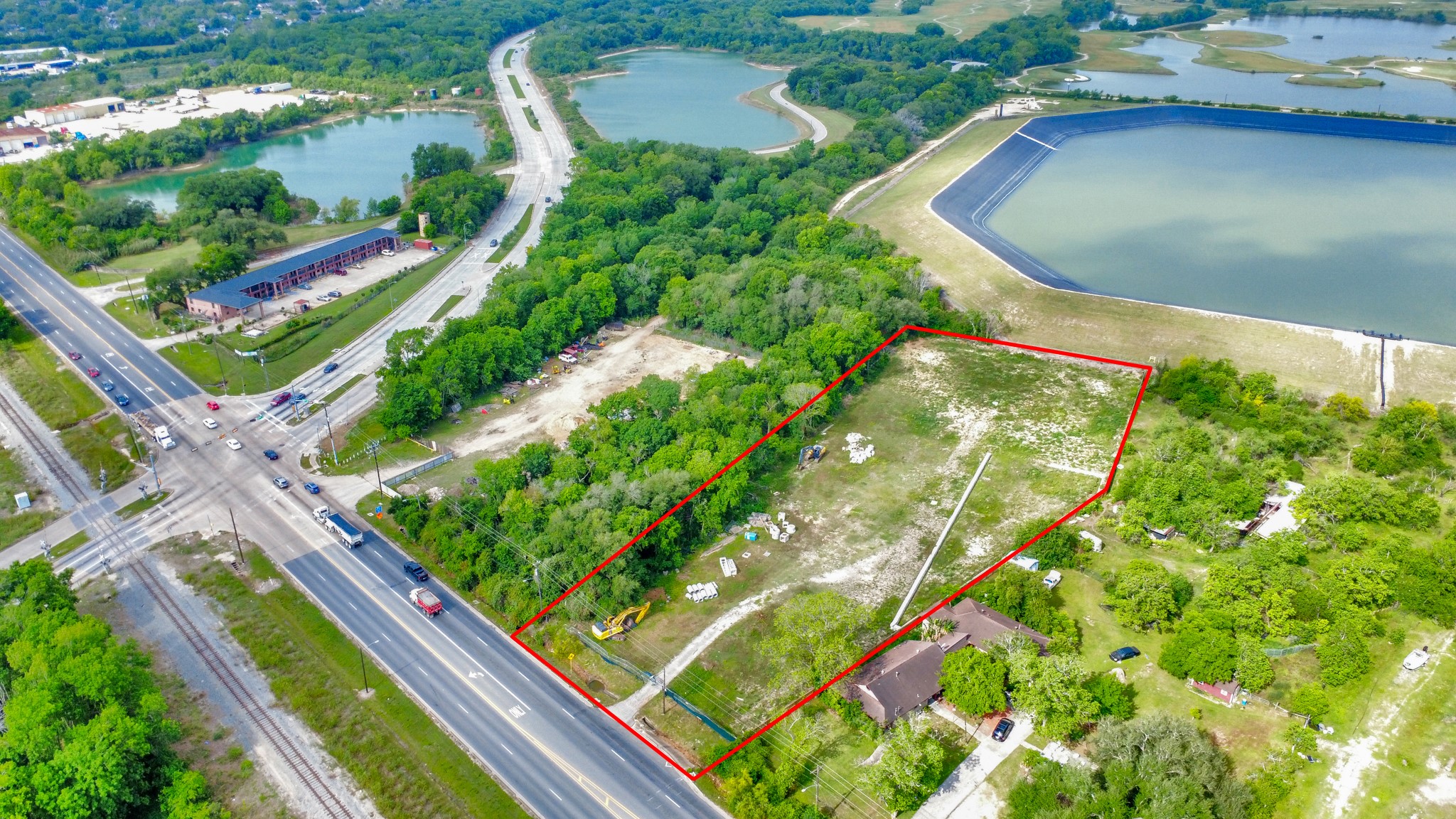 This aerial photo shows a rectangular plot of land outlined in red, bordered by greenery and roads. Nearby are water bodies, a busy intersection, and some buildings, indicating a convenient location with natural surroundings.