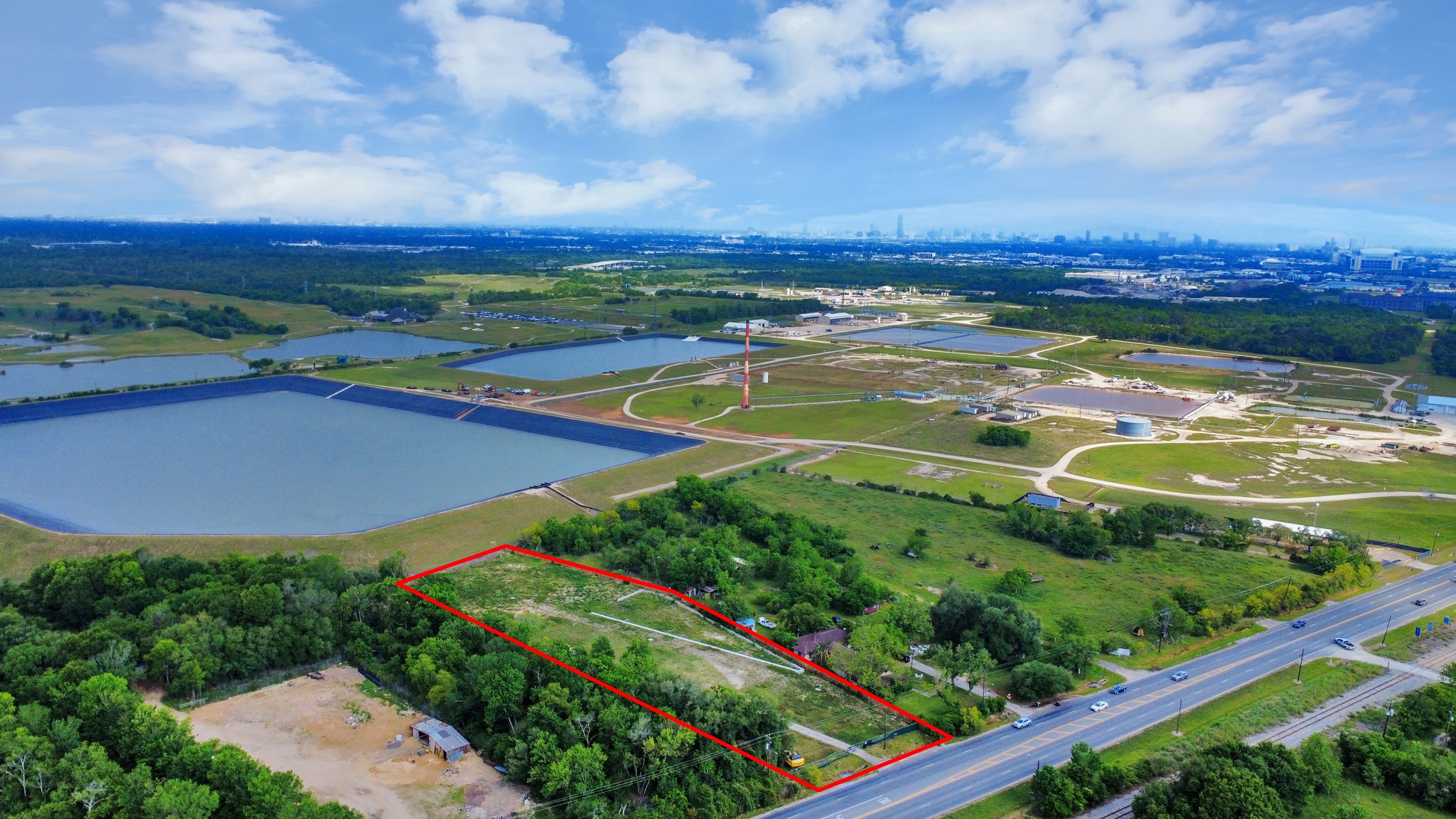 This aerial photo showcases a large plot of land outlined in red, situated near a major road. The area is surrounded by greenery and open fields, with industrial facilities and water bodies visible in the distance. The skyline of a city is faintly visible on the horizon, suggesting a mix of rural and urban proximity.