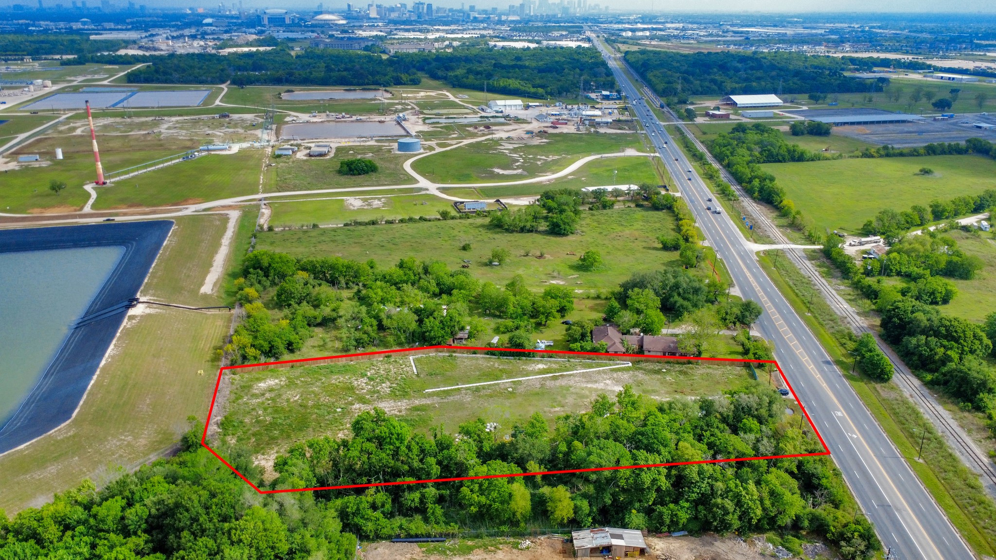 This aerial photo showcases a spacious, rectangular plot of land outlined in red, bordered by greenery. It's situated near a major road with easy access to urban amenities. The surrounding area features open fields and some industrial facilities, with the city skyline visible in the distance.