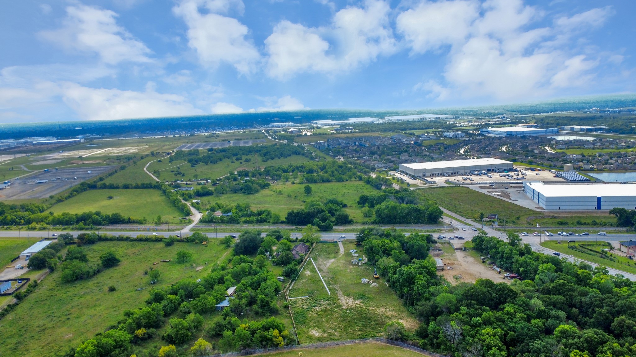 This aerial view showcases a vast, green landscape with scattered trees and open land, bordered by roads. In the distance, there are industrial buildings and a residential area, indicating a blend of rural and urban settings. Ideal for buyers seeking space with nearby amenities.