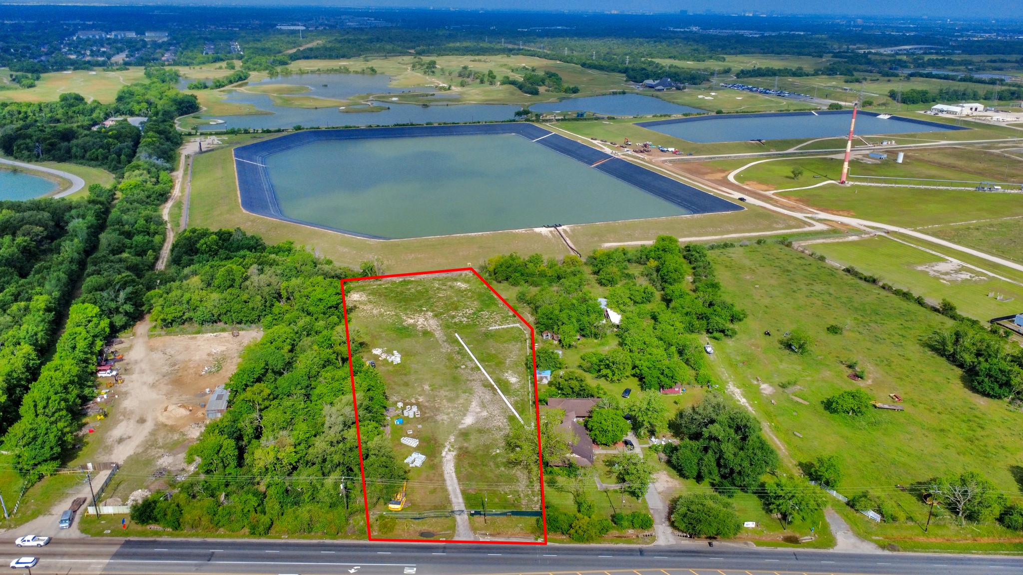This aerial photo shows a large, rectangular plot of land outlined in red, situated next to a road. The surrounding area includes greenery, open fields, and several large water bodies, offering a spacious and scenic environment.