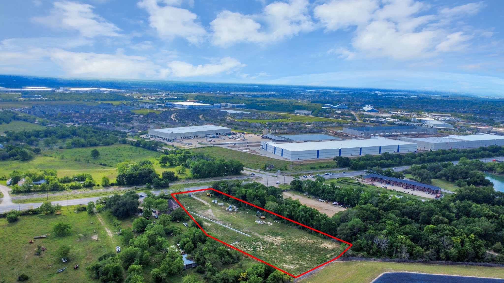 This aerial photo showcases a plot of land outlined in red, situated in a semi-rural area with industrial buildings nearby. The property is surrounded by greenery and has road access, offering a blend of natural and developed surroundings.