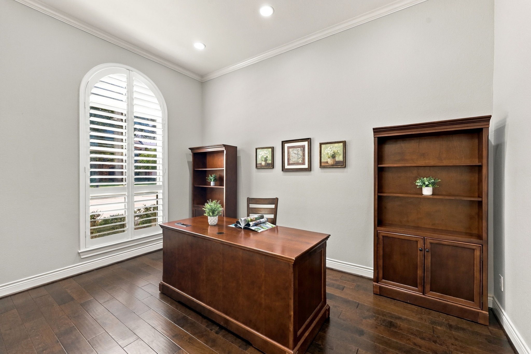 The Home Office features wide-plank wood flooring, an 11-foot ceiling, LED lighting, elegant crown molding, and 5-inch baseboards. A large arched window with plantation shutters adds architectural charm while allowing natural light to fill the space, creating a comfortable and private setting for working from home. The Home Office features wide-plank wood flooring, an 11-foot ceiling, LED lighting, elegant crown molding, and 5-inch baseboards. A large arched window with plantation shutters adds architectural charm while allowing natural light to fill the space, creating a comfortable and private setting for working from home.