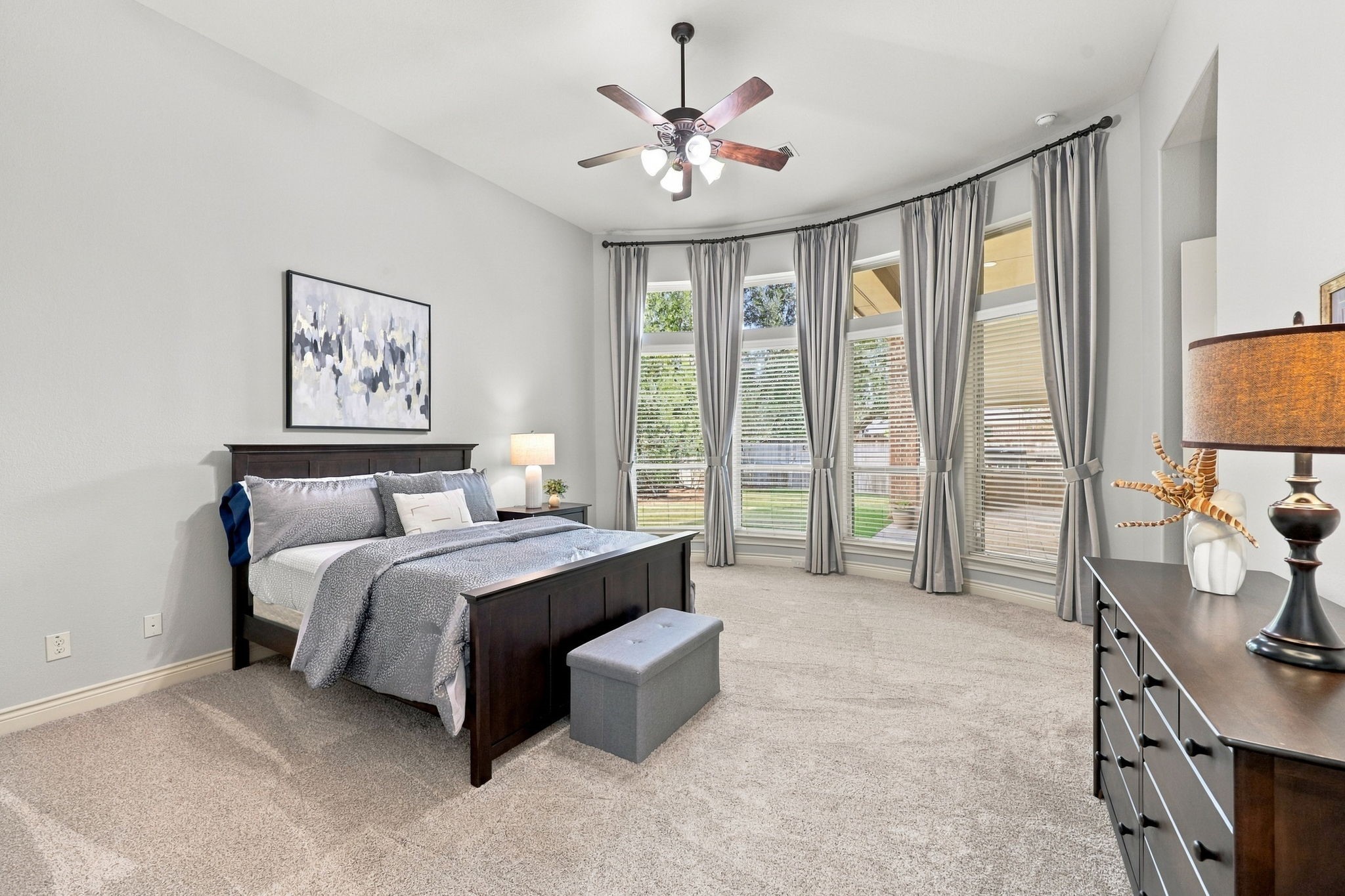 A wall of windows dressed with custom draperies and 2-inch blinds offers a lovely view of the covered back Patio, allowing natural light to flow into the Primary Suite while providing privacy and light control. A 5-blade ceiling fan adds comfort, completing this relaxing private retreat.