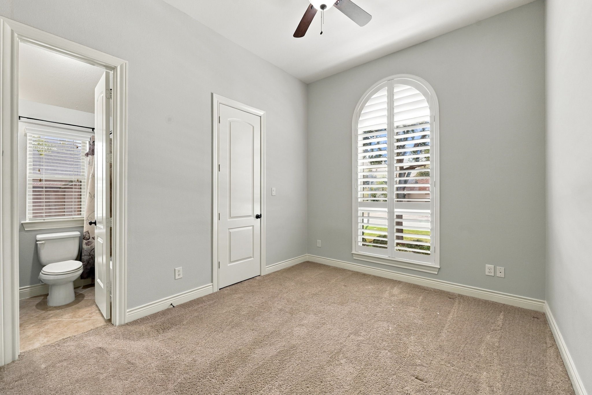 All Secondary Bedrooms are generously sized and feature walk-in closets and direct access to a Bathroom. This Bedroom is located at the front of the home and features a large arched window with plantation shutters, soft, light-colored walls, and wall-to-wall carpeting, creating a bright and comfortable space.