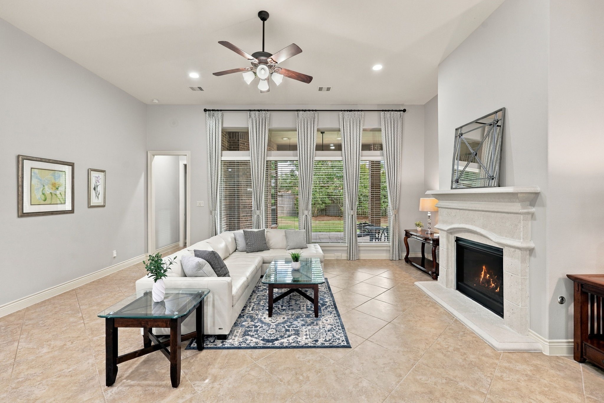 The inviting Family Room features 10-foot ceilings and a beautiful wall of windows overlooking the covered back Patio. Custom draperies paired with 2-inch blinds add a polished touch while providing light control and privacy. A five-blade ceiling fan enhances comfort, while the cast-iron remote-controlled gas-log fireplace creates a warm and welcoming focal point, perfect for relaxing evenings and gathering with family and friends. The doorway to the left leads to the Primary Suite.