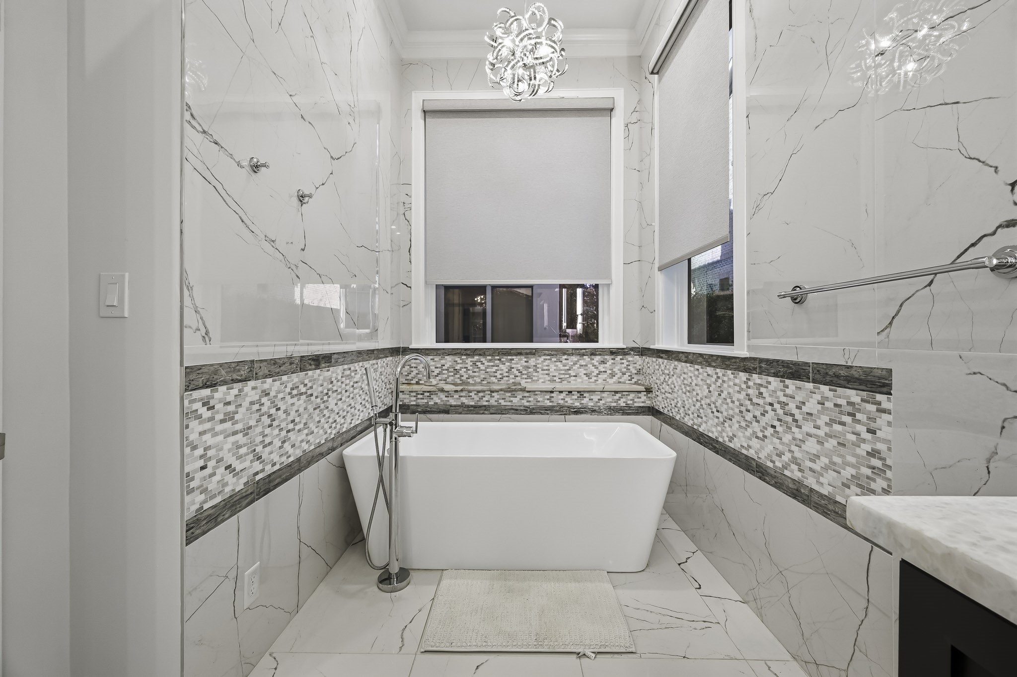 Luxurious bathroom featuring a sleek freestanding tub, and chic mosaic tile accents, all illuminated by a stunning modern chandelier.