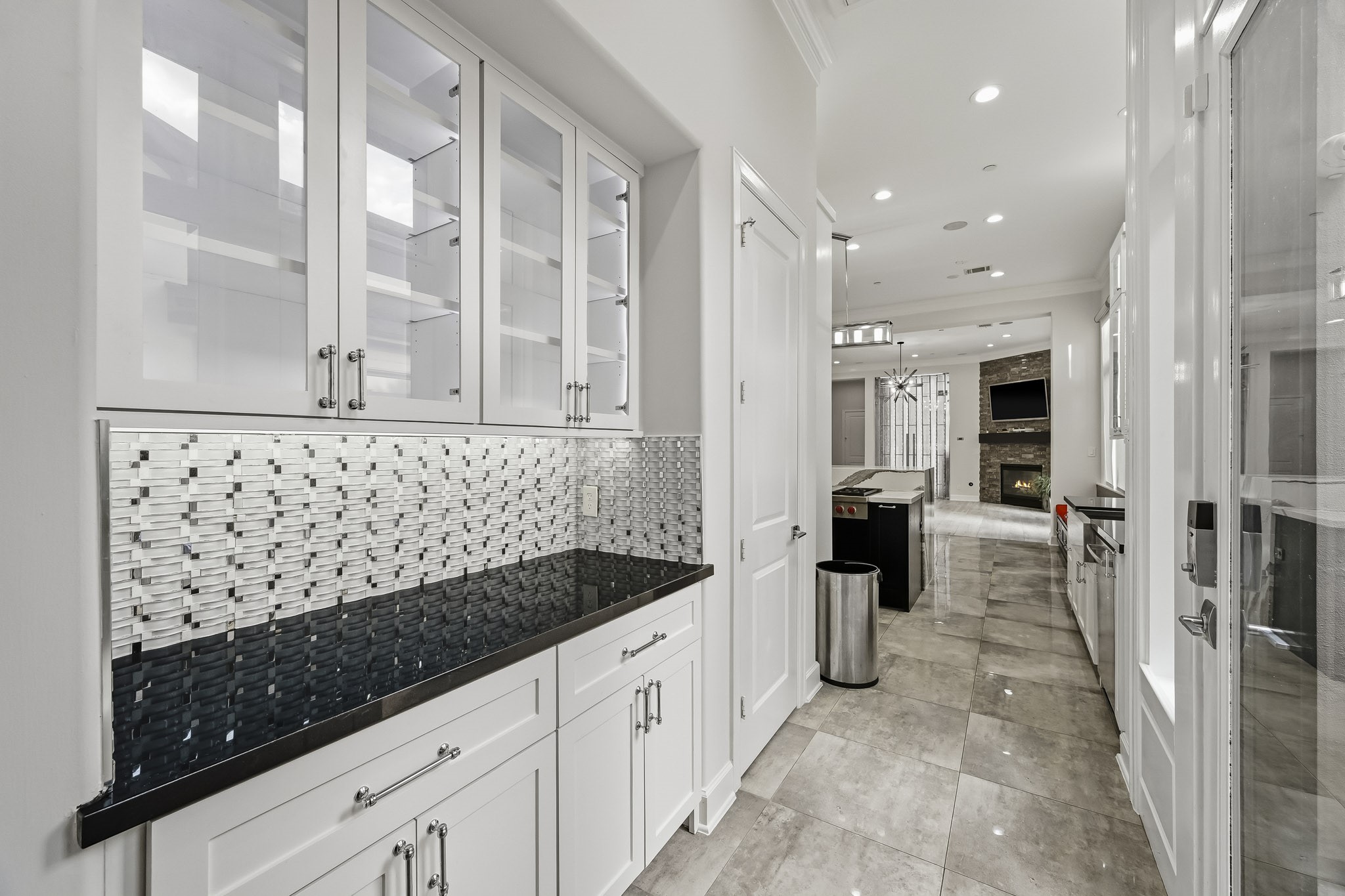 Elegant kitchen hallway featuring sleek black countertops and white cabinetry, highlighted by a modern mosaic backsplash. The open layout leads to a spacious living area with a cozy fireplace.