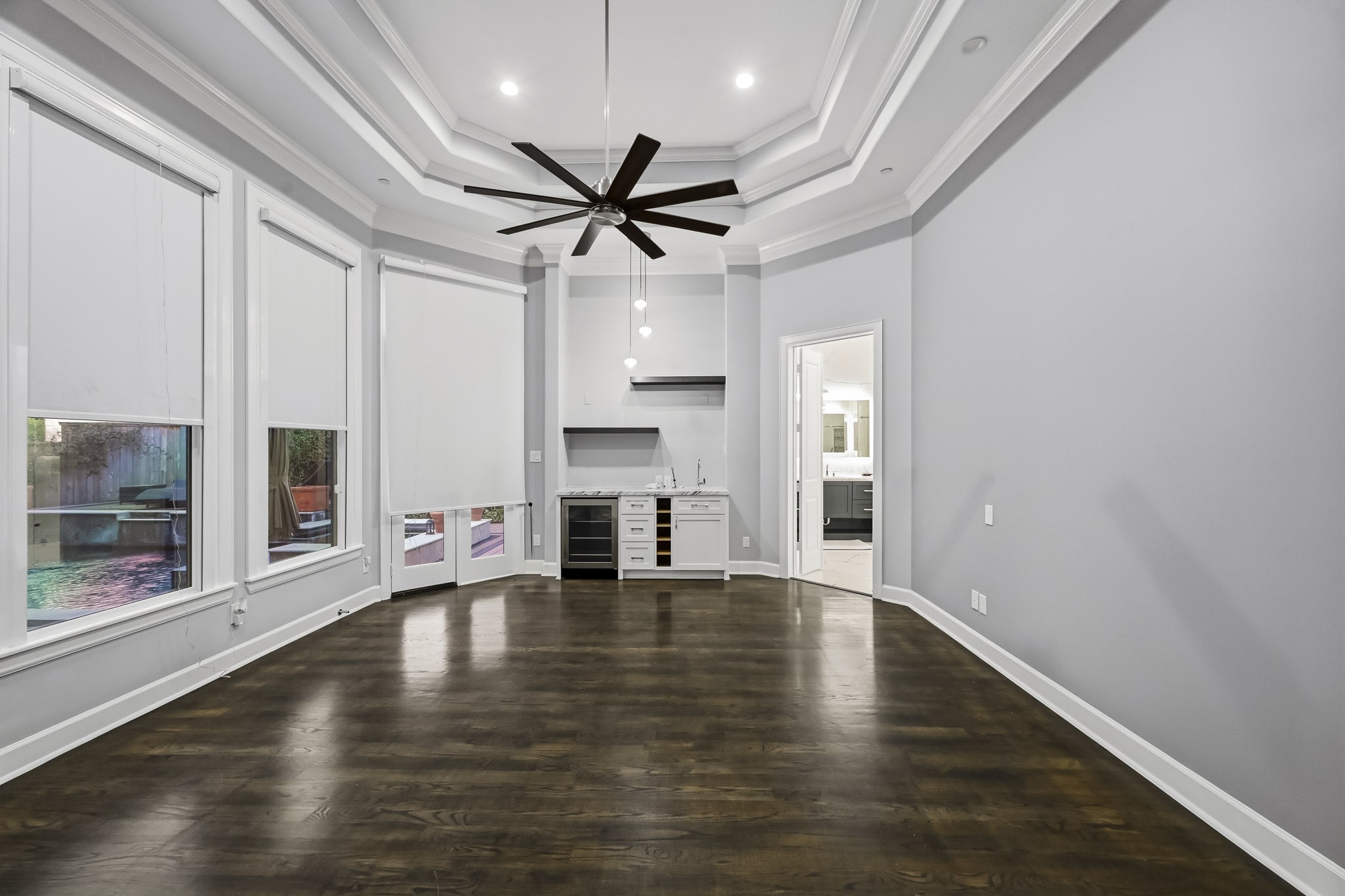 Elegant primary bed room featuring rich floors, a stylish tray ceiling with a modern fan, and ample natural light from large windows with shades. A convenient wet bar adds functionality, while the open layout provides seamless access to an adjacent space.