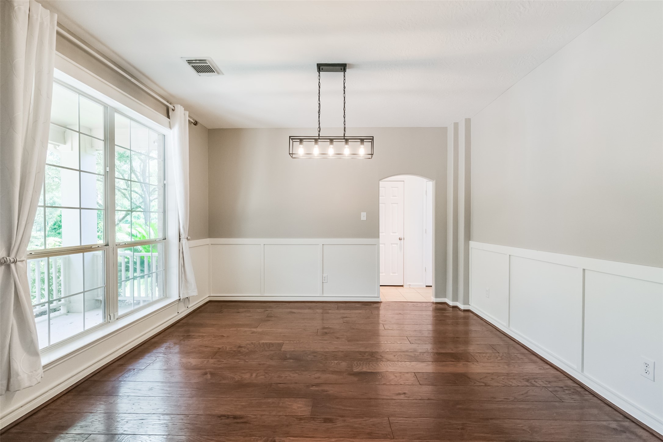 This bright dining room features large windows with white curtains, wood flooring, neutral walls with wainscoting, and a modern light fixture, offering a welcoming and elegant space.