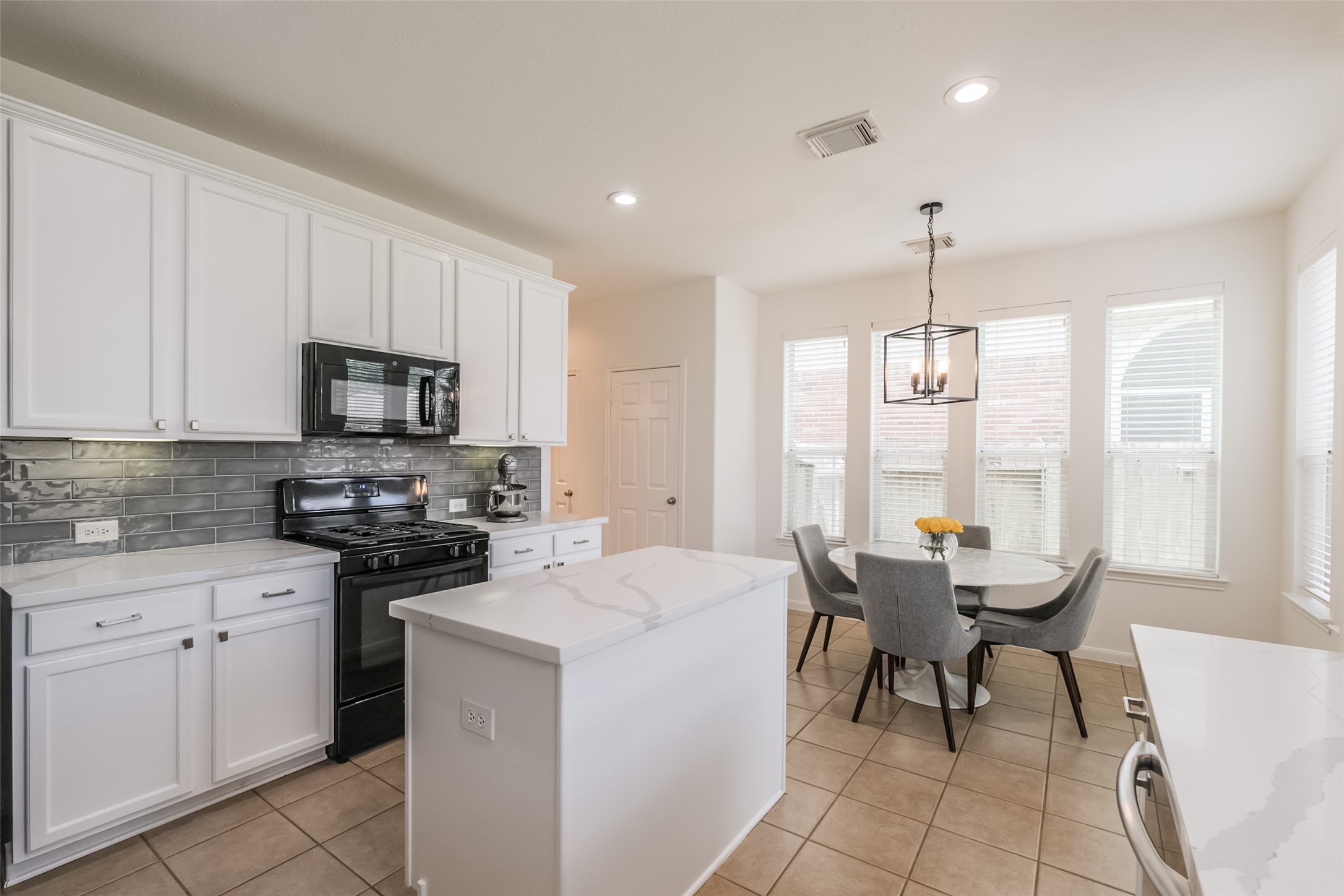 This modern kitchen features sleek white cabinetry, a stylish gray backsplash, and a central island with ample countertop space. It includes a black stove and microwave, with a cozy dining area bathed in natural light from large windows. A contemporary light fixture adds a touch of elegance.