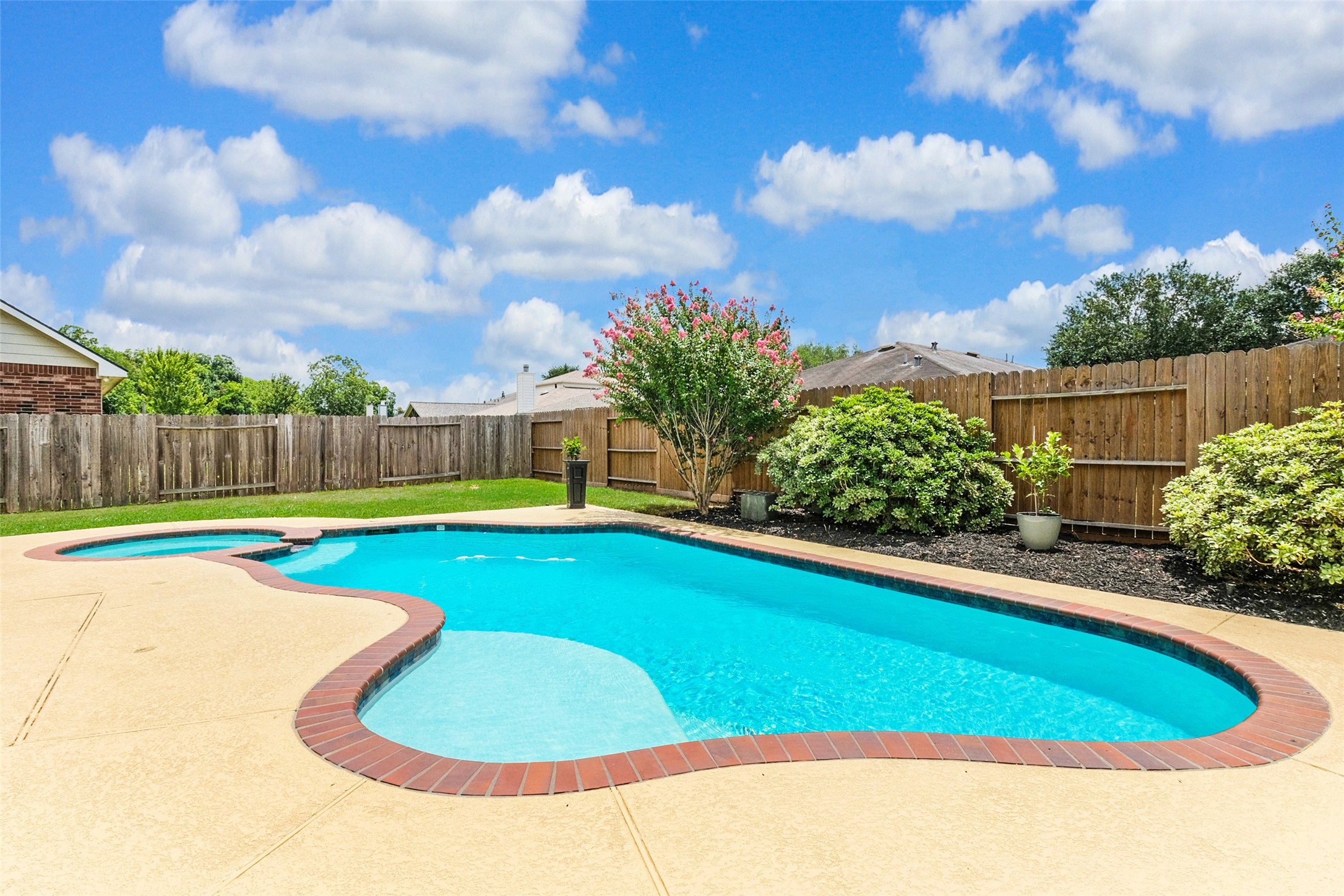 This photo shows a backyard with a well-maintained, curvy swimming pool surrounded by a concrete deck. The yard is enclosed by a wooden fence and features neatly landscaped greenery, creating a private and inviting outdoor space.