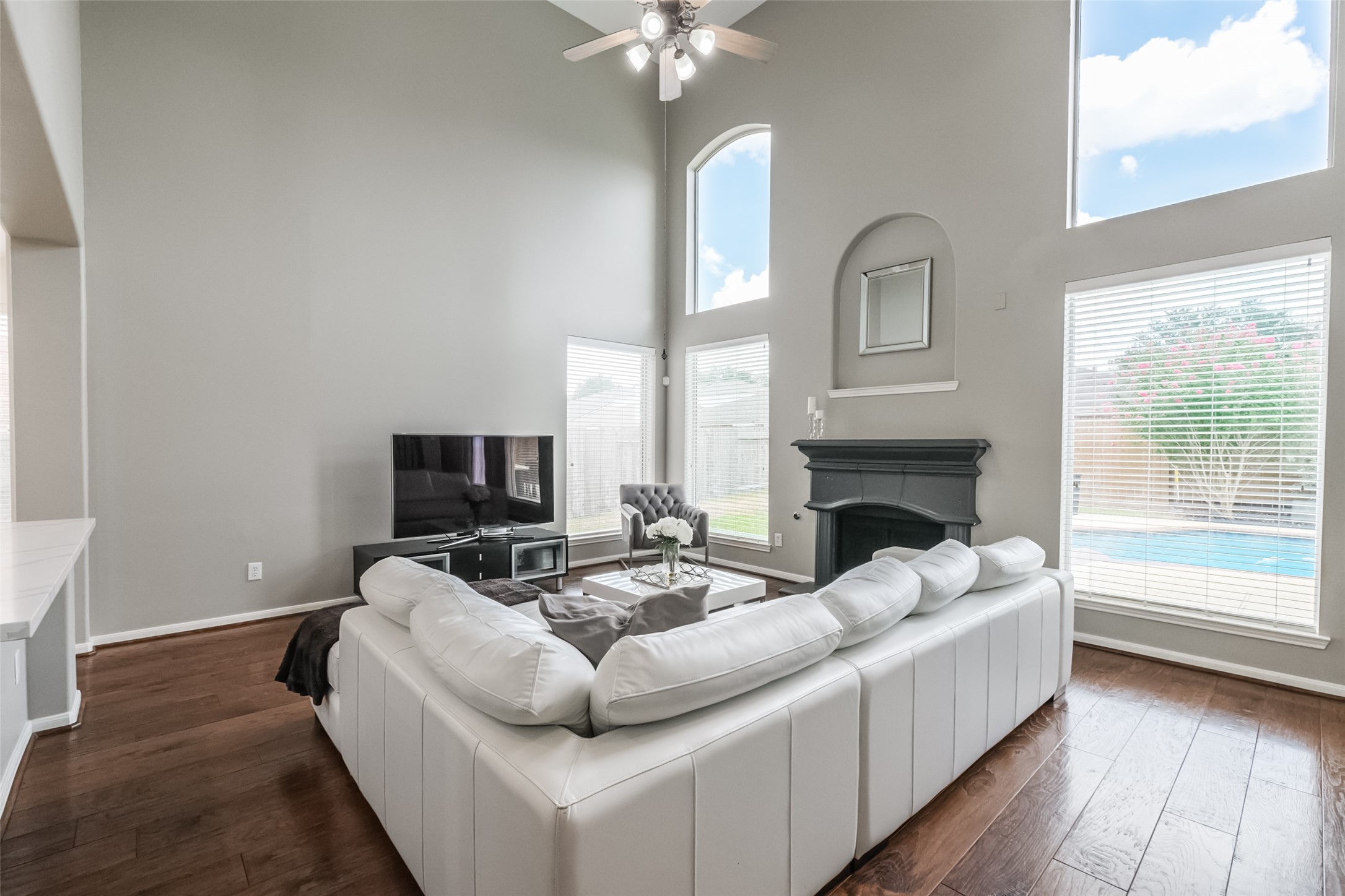 This spacious living room features high ceilings, hardwood floors, and large windows offering plenty of natural light. It includes a cozy fireplace and a view of the pool outside, creating an inviting atmosphere for relaxation and entertainment.