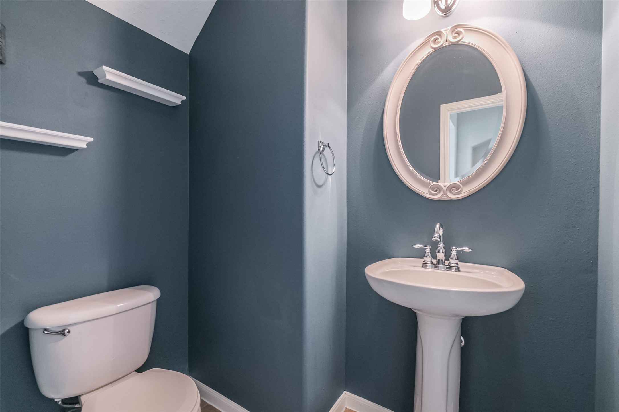 Compact half-bathroom with stylish gray-blue walls, featuring a pedestal sink, oval mirror, and modern light fixture. Practical shelving and a sleek design complete the space.