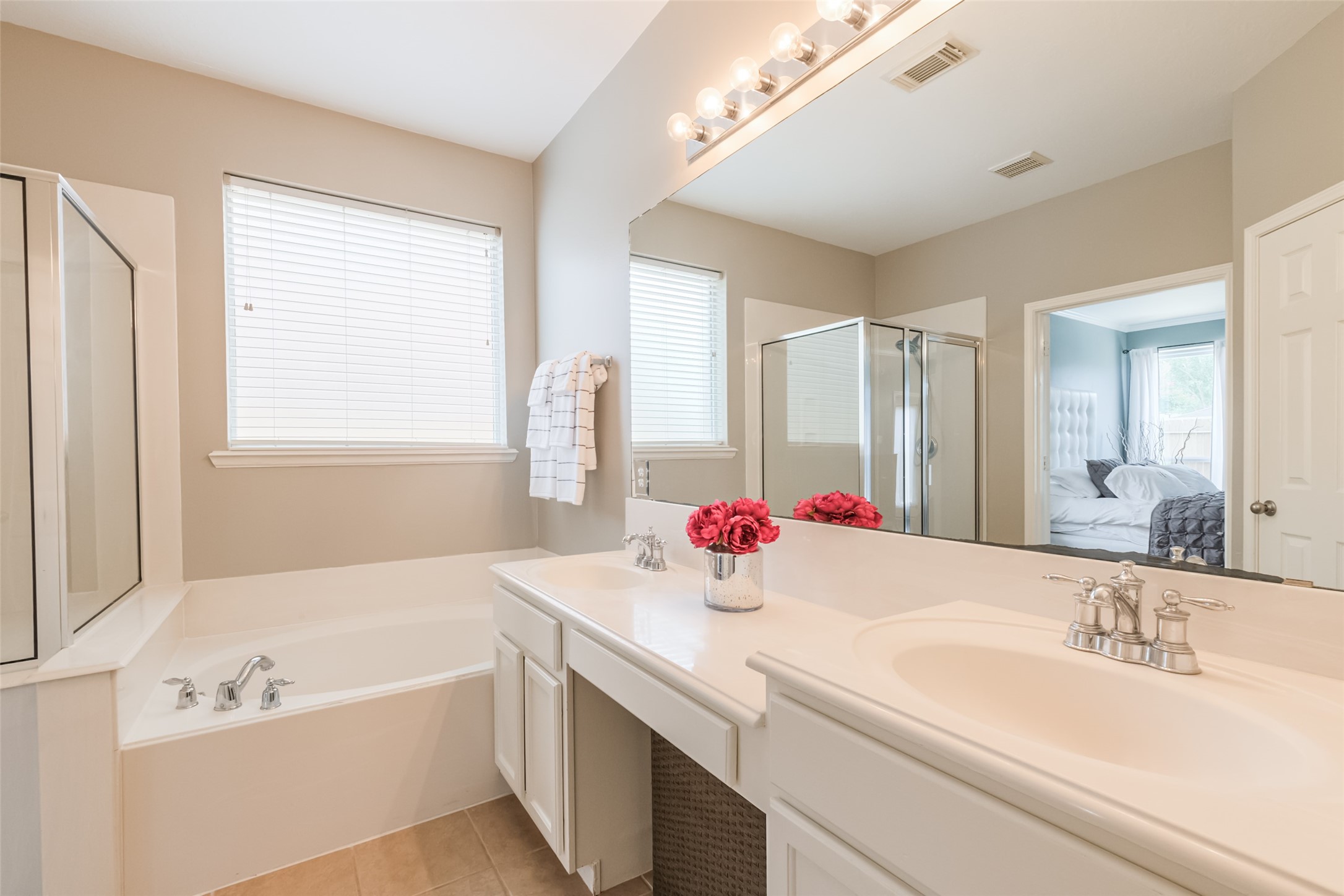 This bright bathroom features a spacious vanity with dual sinks, a large mirror with vanity lights, a soaking tub, and a separate walk-in shower. Neutral tones and natural light create a relaxing atmosphere, with a glimpse into the adjoining bedroom.