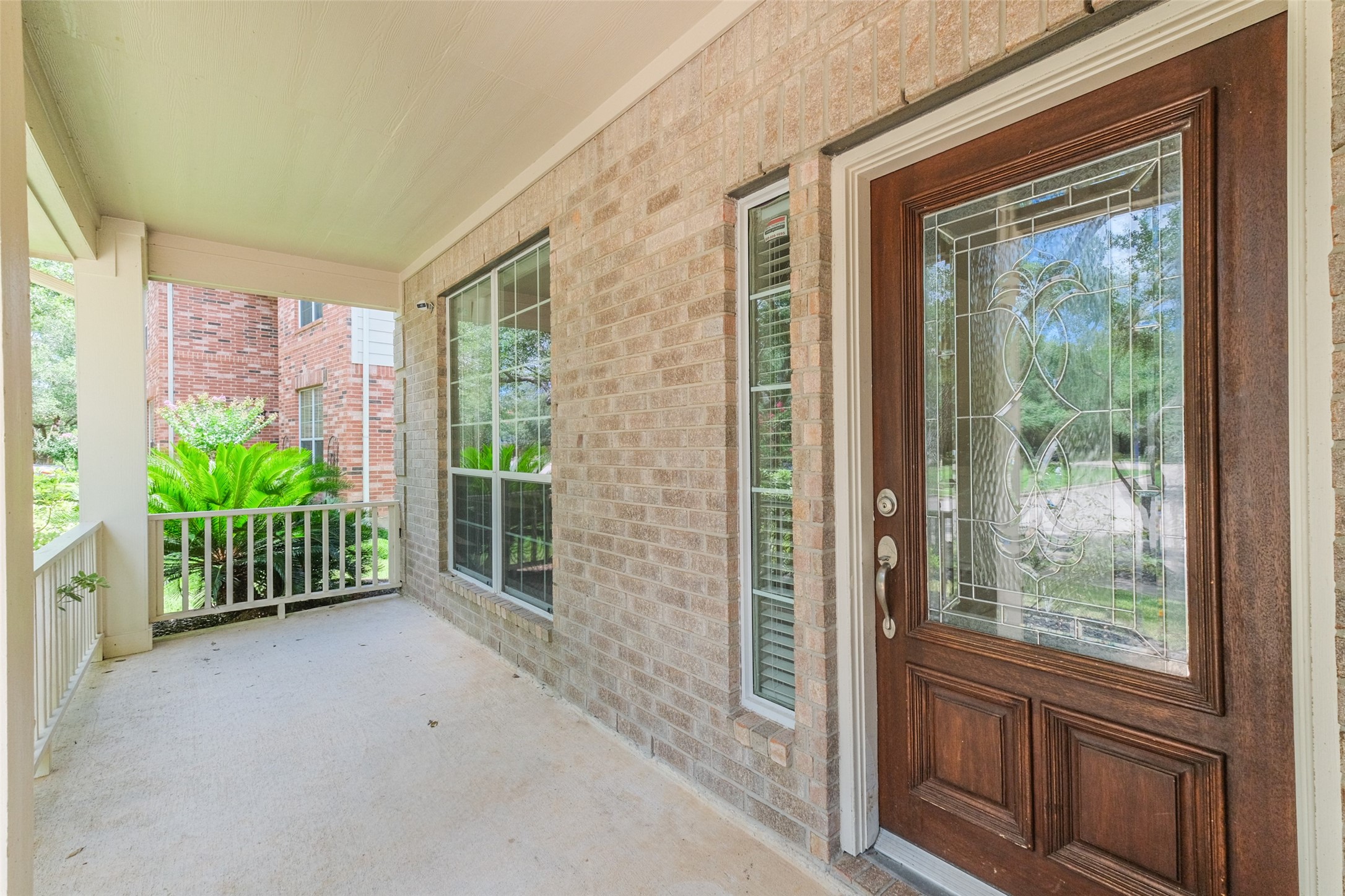 A welcoming front porch with a brick exterior features a decorative glass front door and large windows. The space is sheltered and overlooks a small garden with lush greenery, providing a charming entrance to the home.