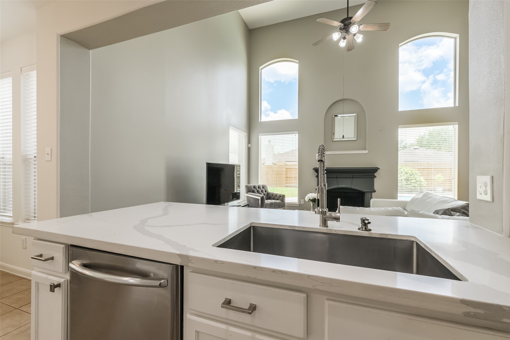 This photo showcases a modern open-concept living area with high ceilings and large windows, providing plenty of natural light. The view from the kitchen features a sleek countertop with a built-in sink, overlooking a cozy living room with a fireplace and ceiling fan, creating a bright and inviting space.