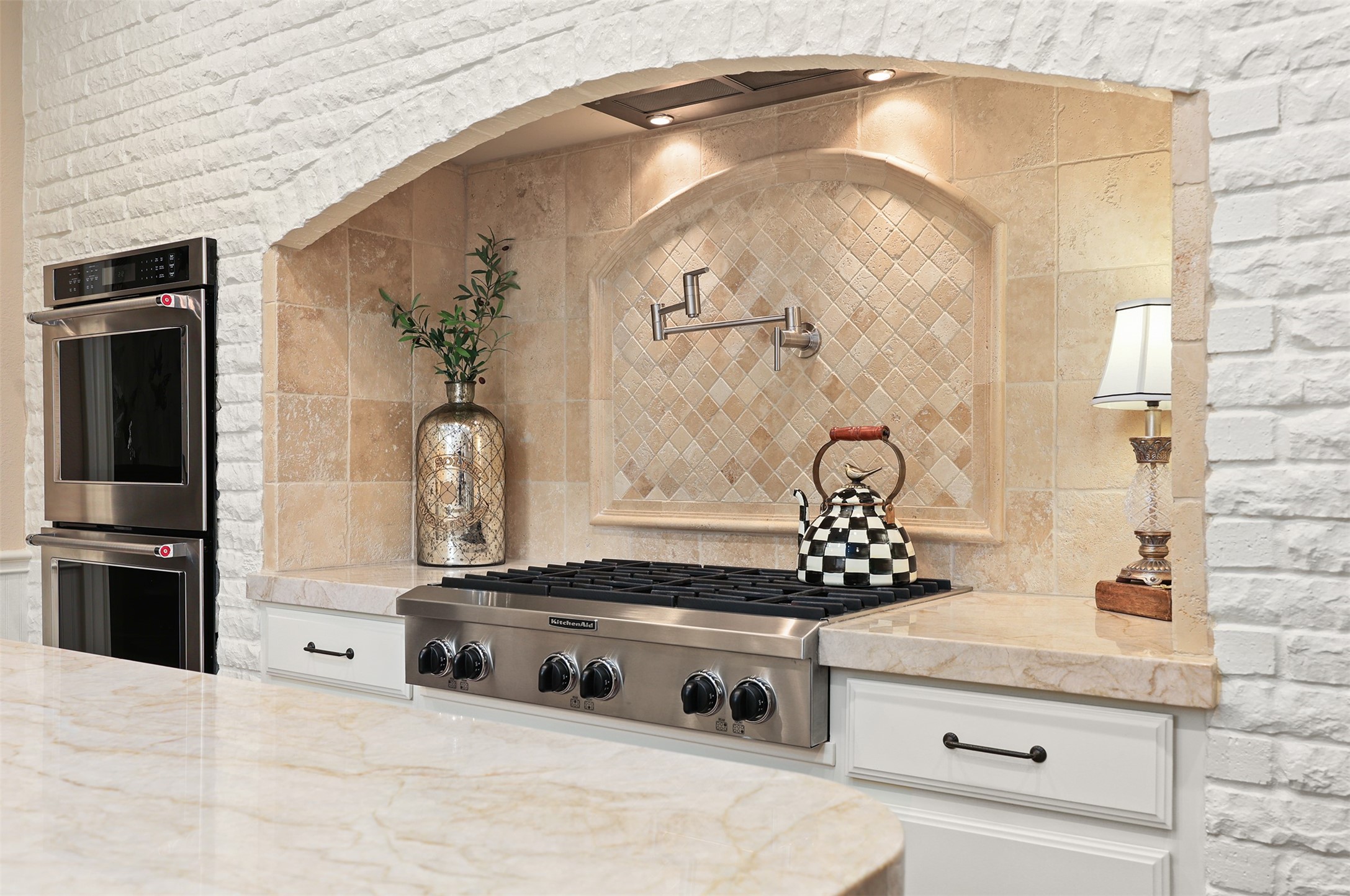 A closer look at the impressive six-burner gas range, framed by a beautiful European-inspired brick archway with custom tilework and a convenient pot filler. Two built-in wall ovens complete this thoughtfully designed cooking space.