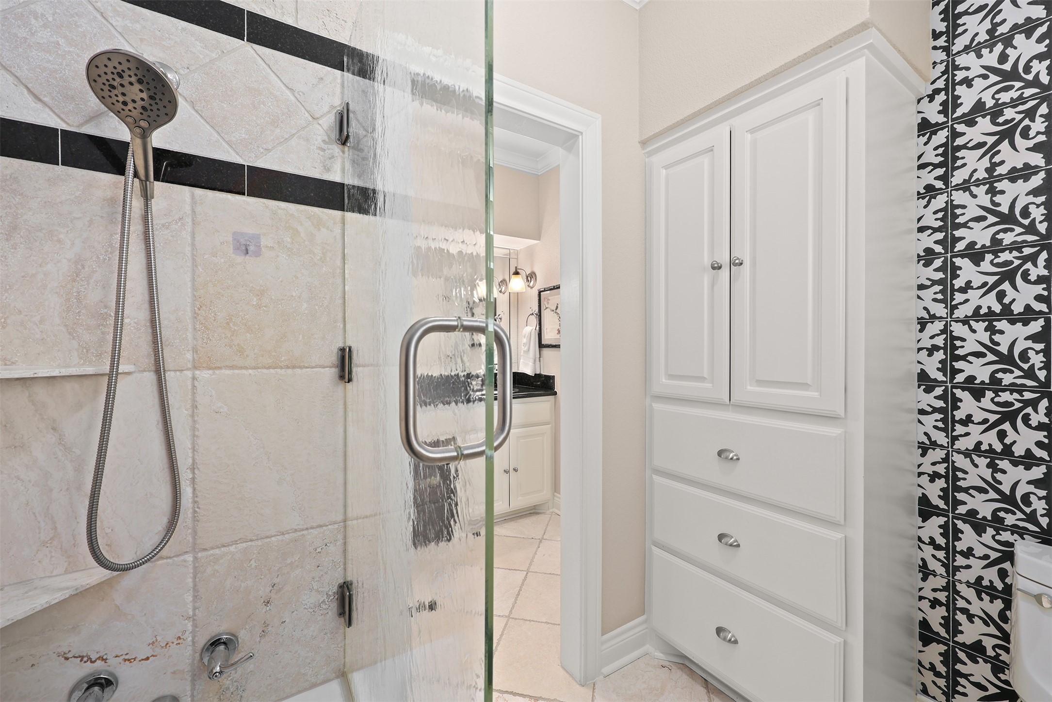 This beautifully designed black-and-white bathroom features abundant custom cabinetry and drawers, a tub/shower combination with tub-to-ceiling tile, and a sleek European-style glass enclosure. Decorative black-and-white patterned tile adds a striking touch of timeless elegance.