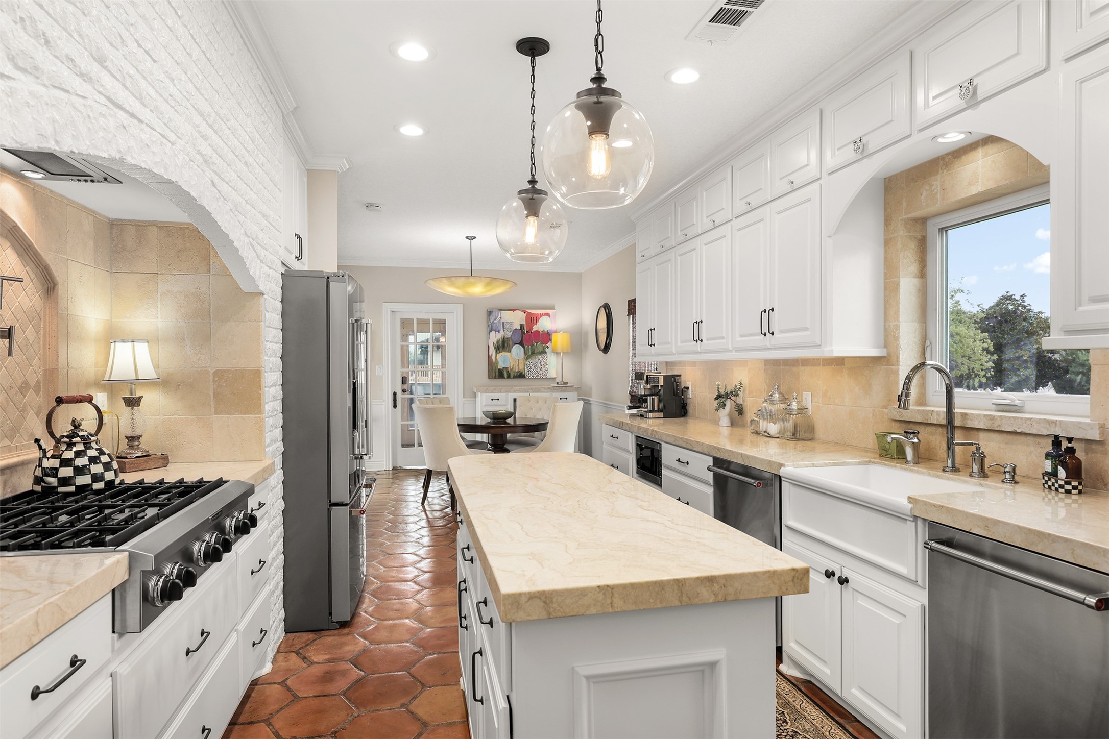 Looking from the wet bar into the beautifully designed kitchen, featuring a spacious center island, stunning Taj Mahal quartzite countertops, an eight-burner gas range, two dishwashers, and a farmhouse sink perfectly positioned beneath a window overlooking the outdoors.
