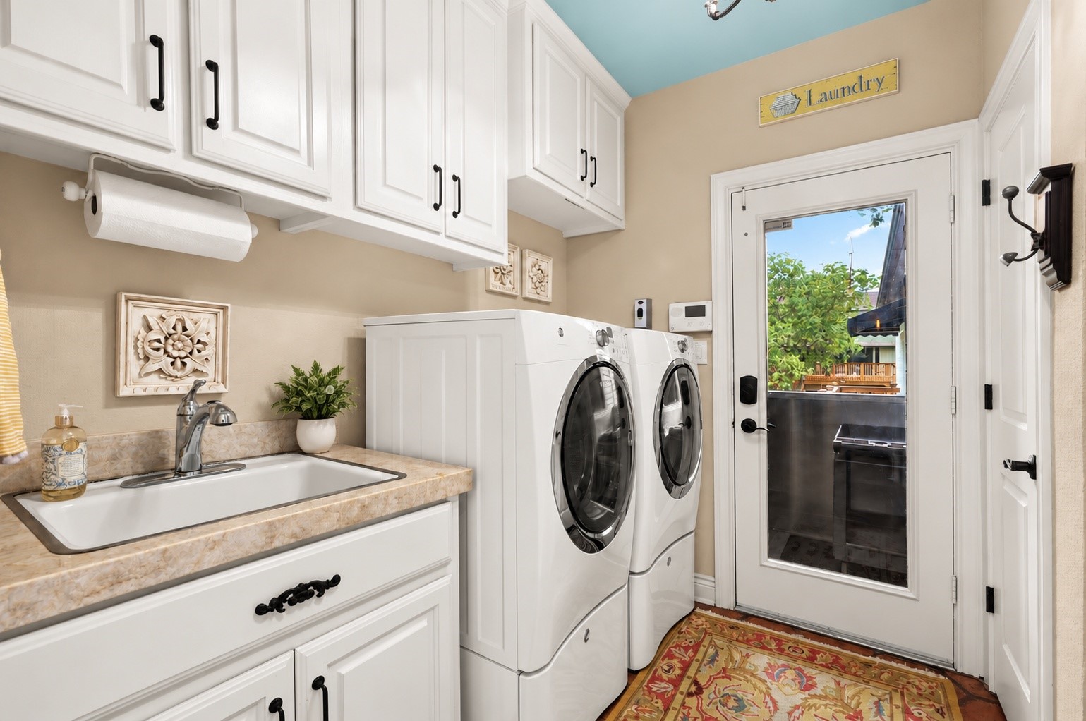 Spacious laundry room featuring abundant storage cabinetry, a utility sink with Taj Mahal quartzite countertop, and a convenient half bath. A glass door leads to the covered exterior elevator/lift, ideally located near the kitchen for easy loading and unloading of groceries. Refrigerator in laundry room conveys with the house.