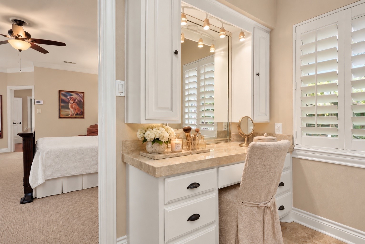 Charming makeup vanity features an elegant Taj Mahal countertop and a nearby window providing beautiful natural light. The primary bath also offers separate his-and-hers walk-in closets, and a spacious linen closet with a convenient built in laundry hamper, providing exceptional storage and organization.