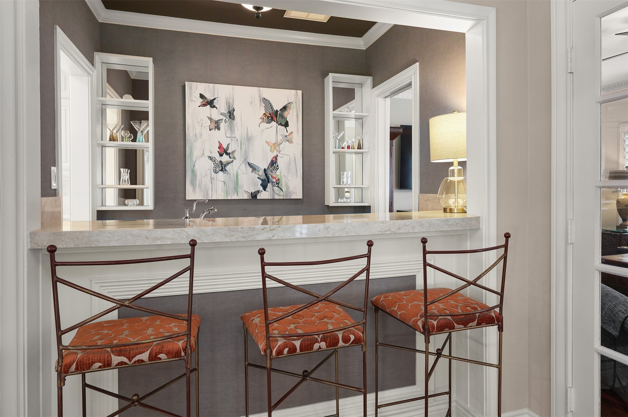 View from the foyer into the beautifully designed pass-through wet bar, featuring a stunning Taj Mahal quartzite countertop and seating for three. The television room is located to the right through the French doors, with the kitchen accessible beyond the bar.