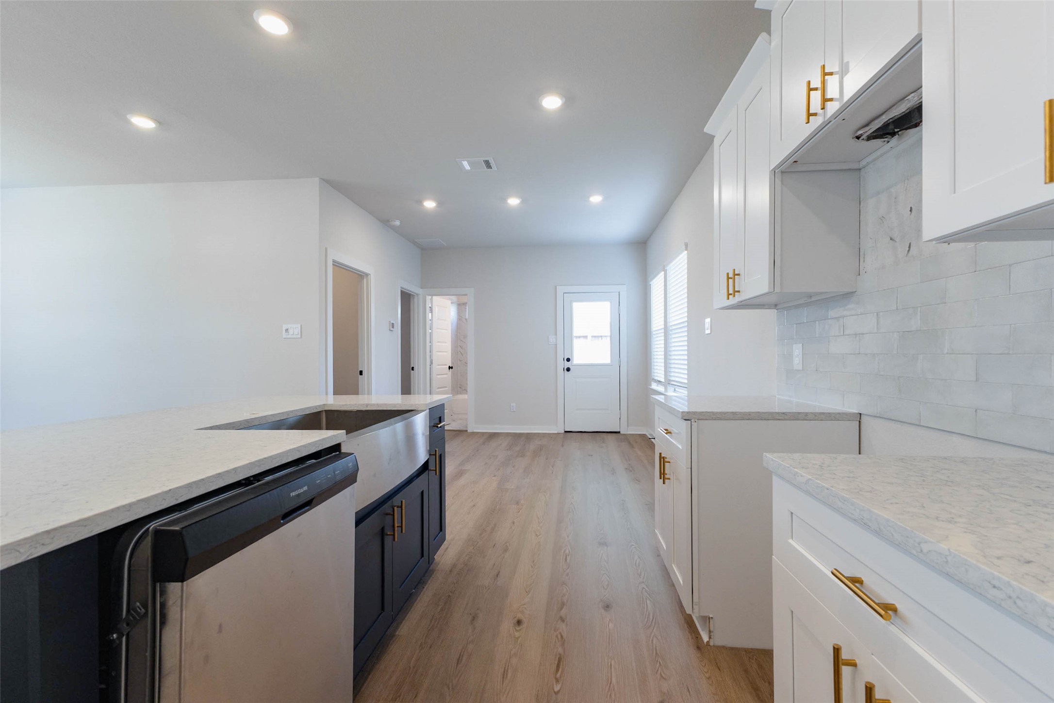 Here is a closer look at the work zone: apron-front sink, stainless dishwasher, marble-look tile backsplash and long quartz counters with plenty of landing space. Gold cabinet pulls add a touch of warmth against the white shaker doors. Beyond the kitchen, a short hallway leads to the back door and secondary rooms, so groceries and everyday traffic move through the home easily.
