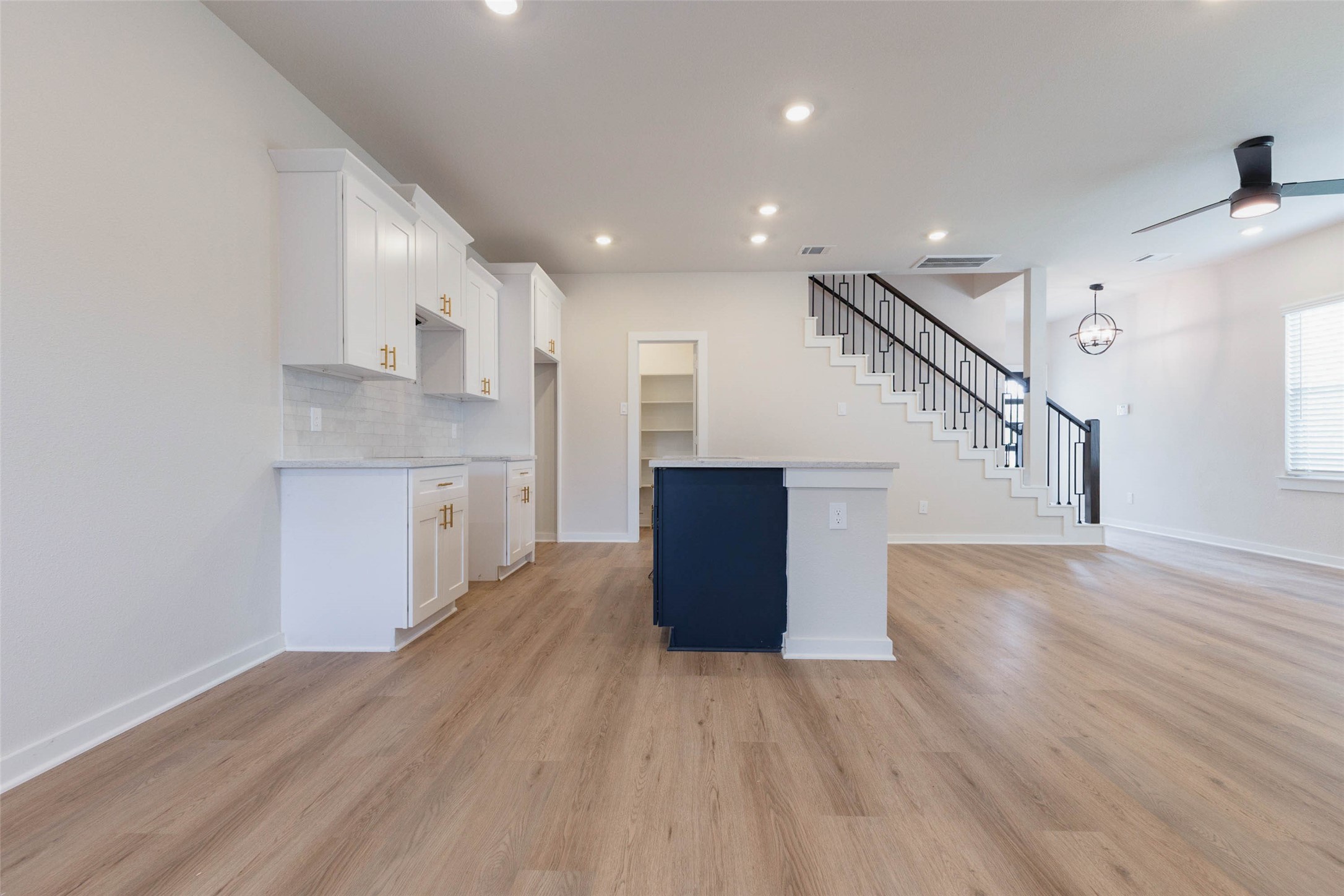 A navy blue island adds a pop of color against crisp white cabinetry and soft gold hardware, giving this kitchen a custom, designer look. The tiled backsplash, deep upper cabinets and walk-in pantry shelving show how much storage is built in. A gas range supports serious home cooking, and the open layout keeps the chef connected to the living room and the staircase landing nearby.