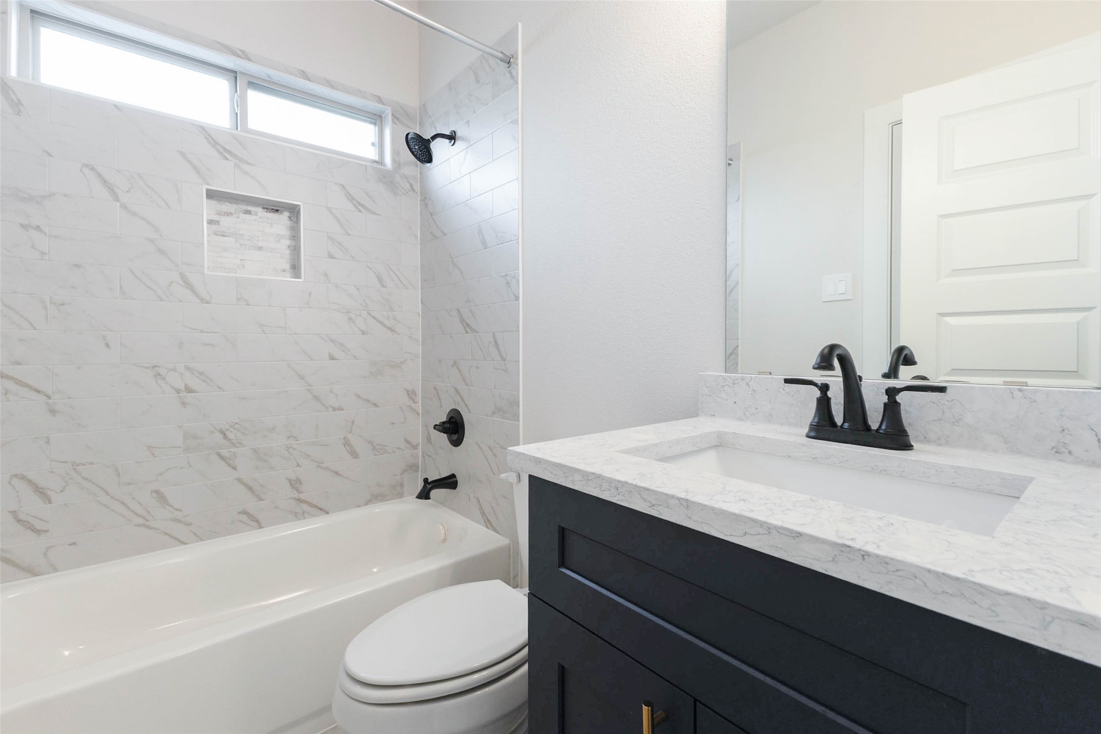 A full bath near the bedroom wing features a tub-shower wrapped in marble-look tile with a built-in niche for shampoo and soap. The navy vanity, quartz top and matte black fixtures create a modern, high-end look, and a transom window above the tub brings in natural light. It is a practical, stylish space for guests, kids or anyone needing a bath close to their bedroom.