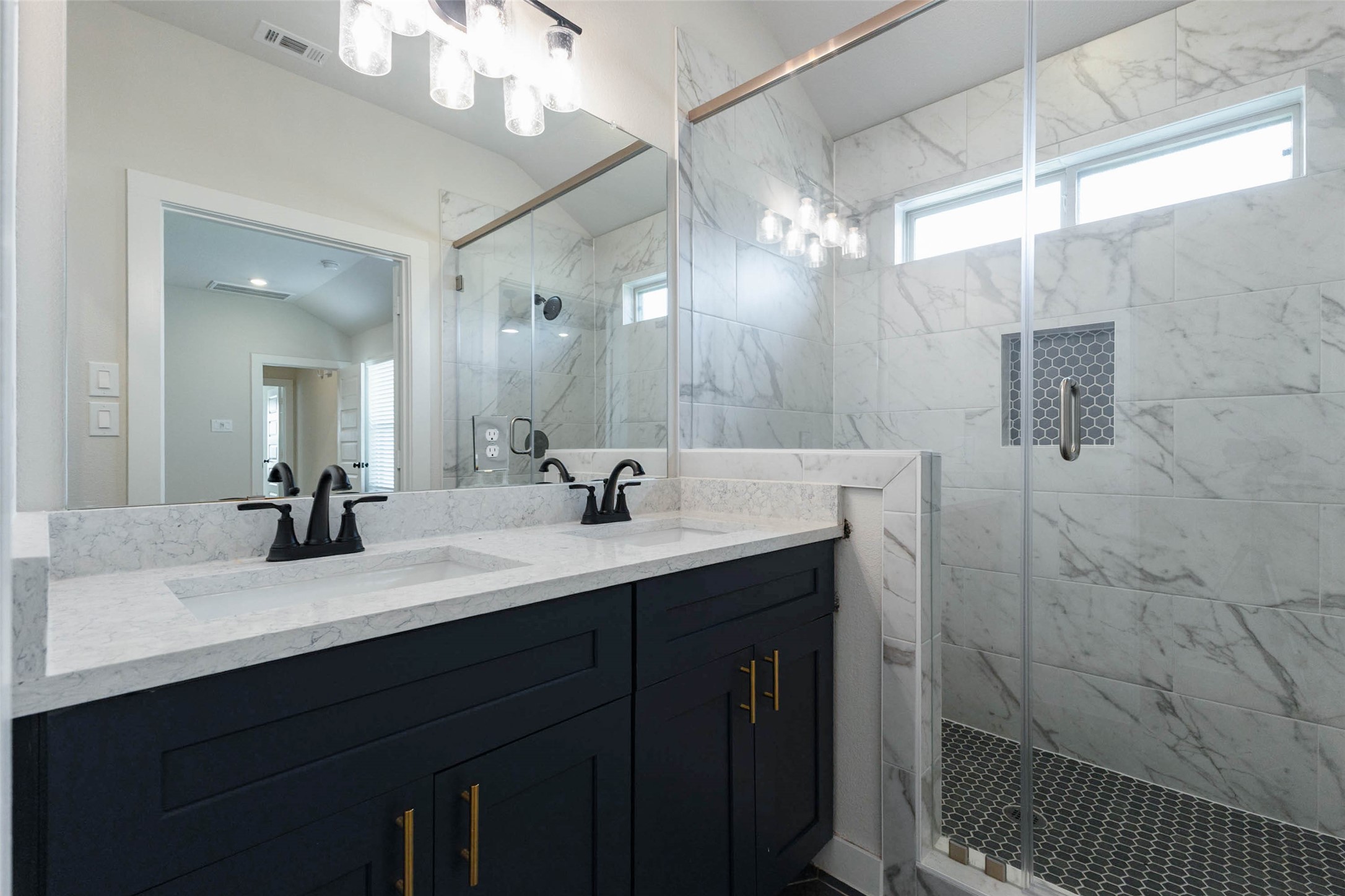 Another view of the primary bath shows the glass-enclosed shower with a built-in niche, hex-tile floor and a high window that brings in daylight while keeping things private. Black faucets contrast beautifully with the white quartz counters and navy vanity cabinets, and the large mirror makes the room feel even bigger. It is a polished, low-maintenance space designed for daily use.