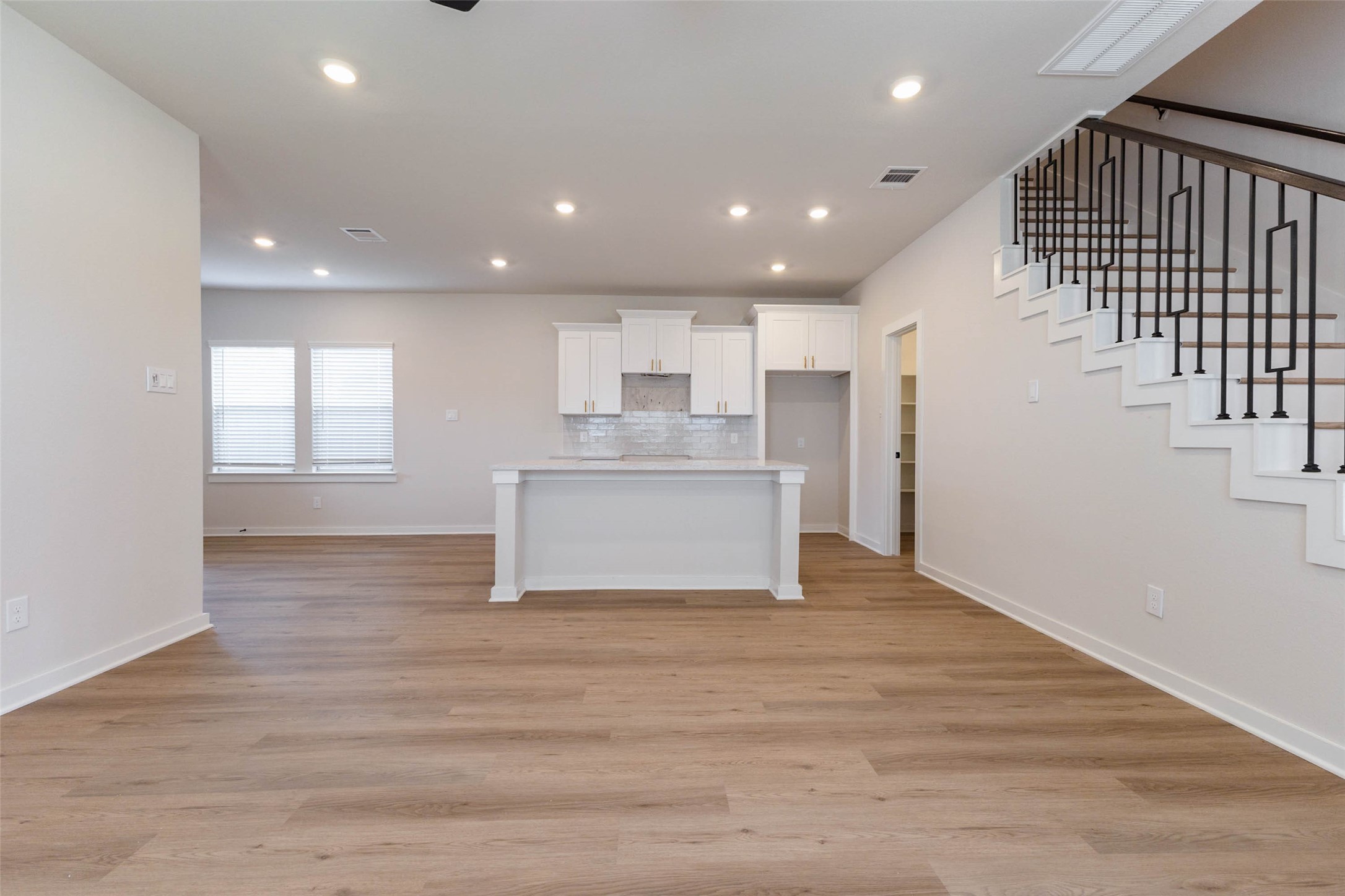 Looking from the living room back toward the kitchen, the quartz-topped island becomes the heart of the home, with space for barstools on one side and prep room on the other. White shaker cabinets reach up high for extra storage, and the staircase and walk-in pantry sit just beyond. The straight sightlines make it easy to keep an eye on the whole main floor while you cook.
