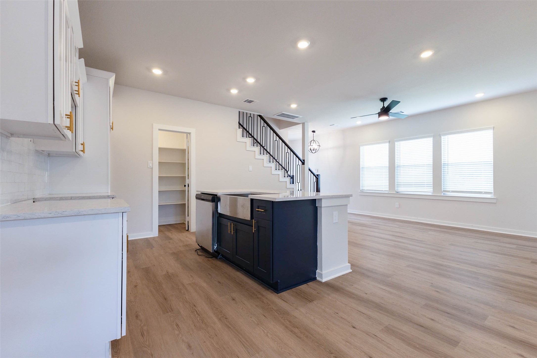 This angle shows the kitchen island paired with a farmhouse-style sink and stainless dishwasher, a combination that is as practical as it is beautiful. The walk-in pantry just behind offers shelving for bulk groceries and small appliances, and the quartz counters wipe clean in seconds. Windows across the back of the room keep the whole space bright, from morning coffee to evening cleanup.