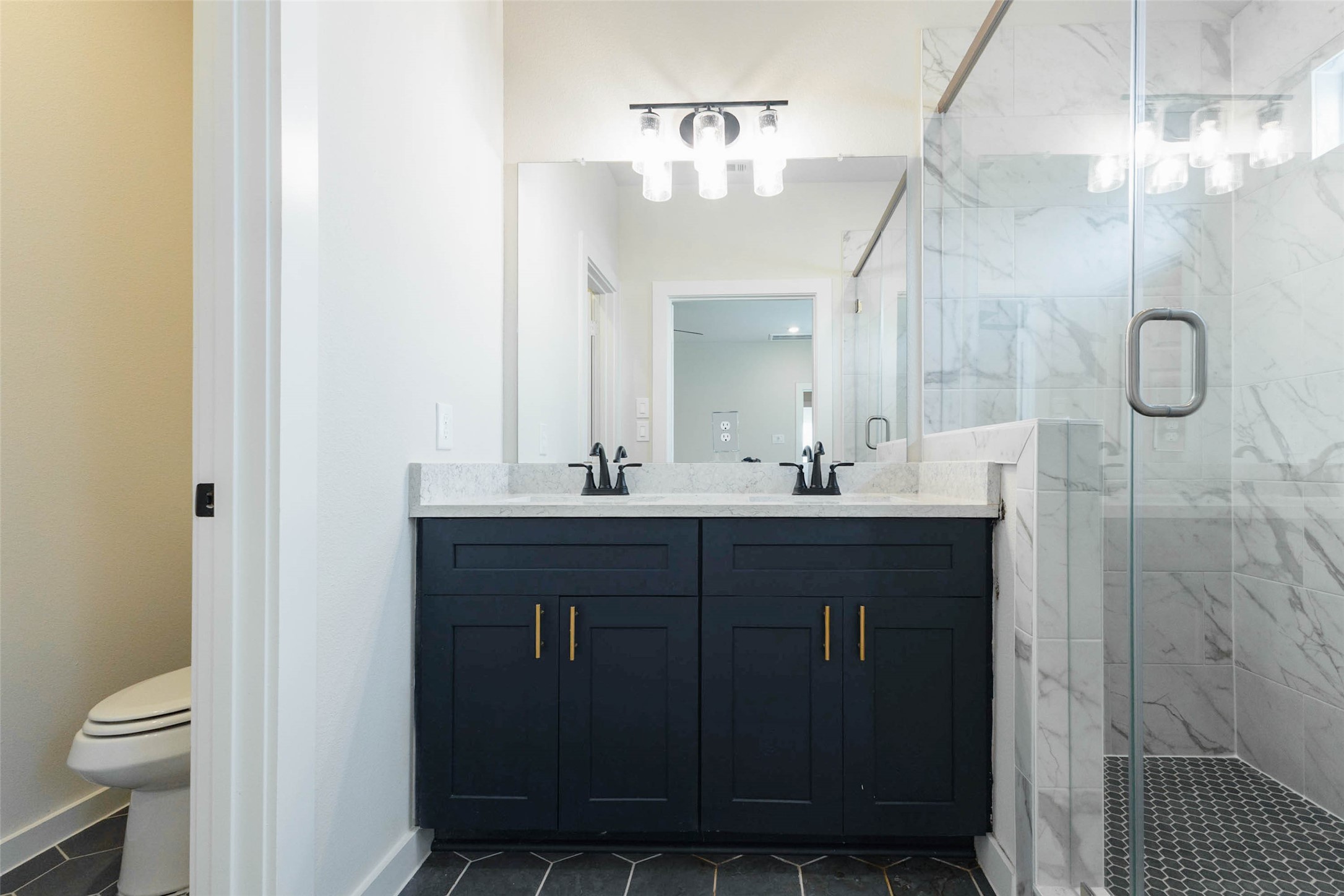 The primary bath is a retreat, with a dual-sink navy vanity, quartz counters, gold pulls and a frameless glass shower wrapped in marble-look tile. A private water closet gives extra privacy, and the stylish light fixture adds a bit of sparkle. Two sinks mean no waiting in the morning, and the dark cabinets against bright tile make it feel like a boutique hotel.