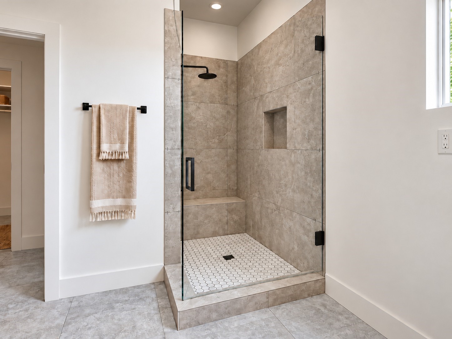 Experience a beautifully designed walk-in shower featuring elegant floor-to-ceiling textured tile, sleek frameless glass doors, and sophisticated matte black fixtures. A rainfall showerhead provides a spa-inspired experience, while the built-in bench and recessed storage niche offer everyday convenience. The white hexagonal mosaic flooring adds a stylish contrast to the warm neutral tile. These photos have been virtually rendered for visual reference. Experience a beautifully designed walk-in shower featuring elegant floor-to-ceiling textured tile, sleek frameless glass doors, and sophisticated matte black fixtures. A rainfall showerhead provides a spa-inspired experience, while the built-in bench and recessed storage niche offer everyday convenience. The white hexagonal mosaic flooring adds a stylish contrast to the warm neutral tile. These photos have been virtually rendered for visual reference.