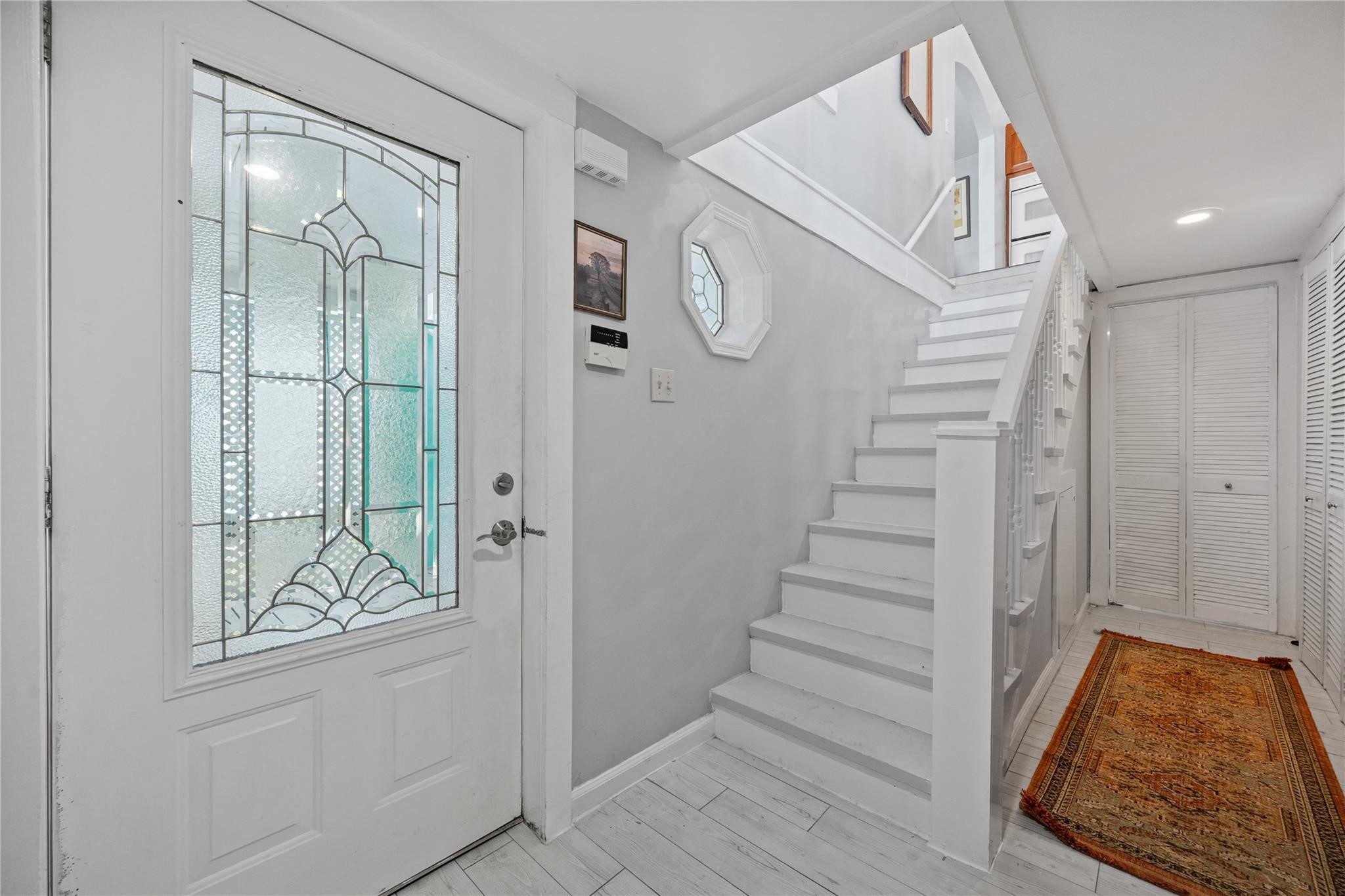 Front Entrance & Staircase: Welcoming front entrance showcasing a beautiful decorative leaded-glass entry door and classic white staircase, highlighting the home's charming architectural character. Wood-look tile flooring and coastal-inspired finishes complement the inviting entryway, with stairs leading to the second-floor kitchen, living and dining areas, and outdoor upper deck.