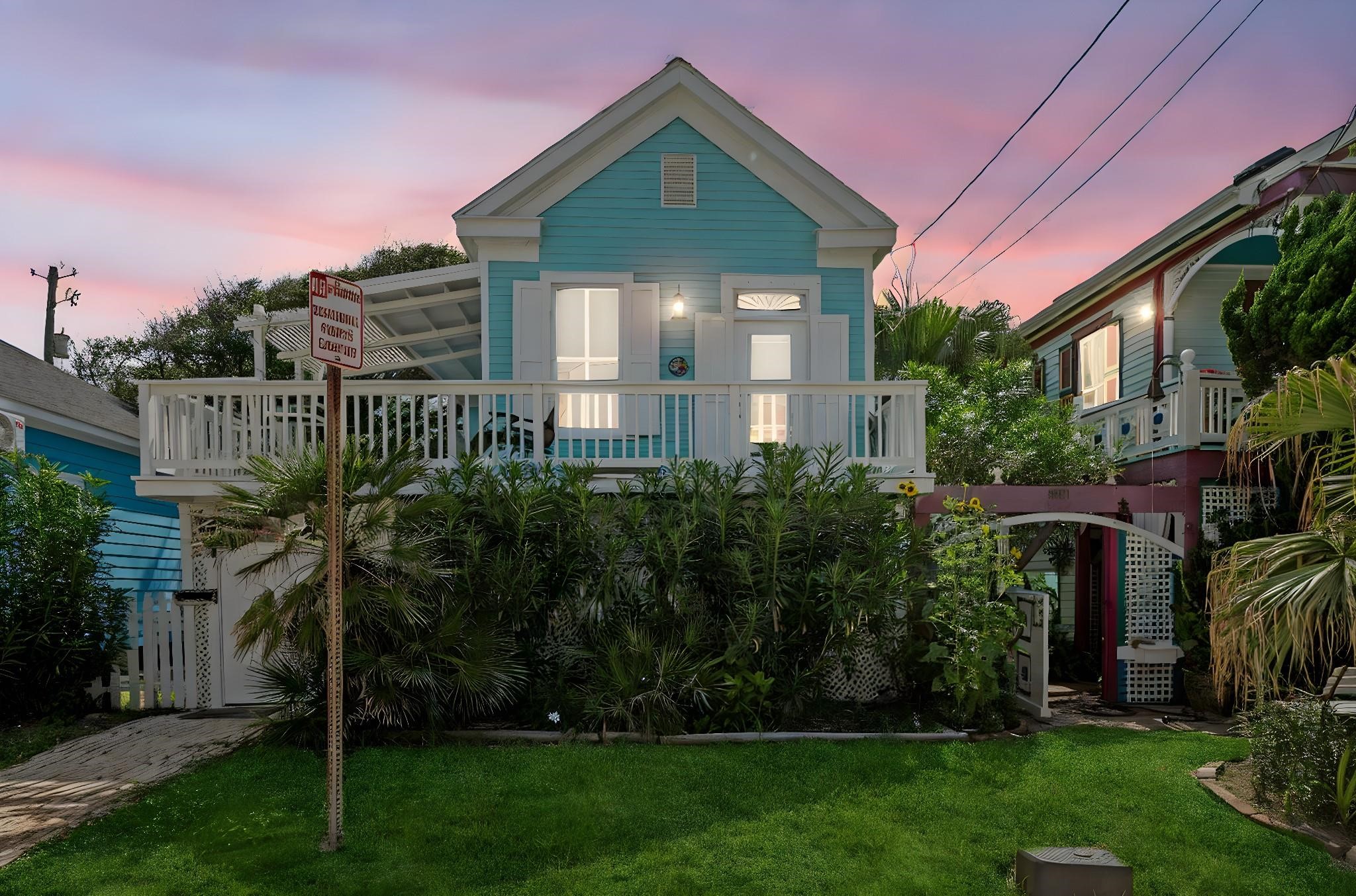 Twilight view of this charming coastal cottage, featuring an elevated front porch, tropical landscaping, and inviting exterior lighting in a prime Galveston location approximately a block and a half from the beach.