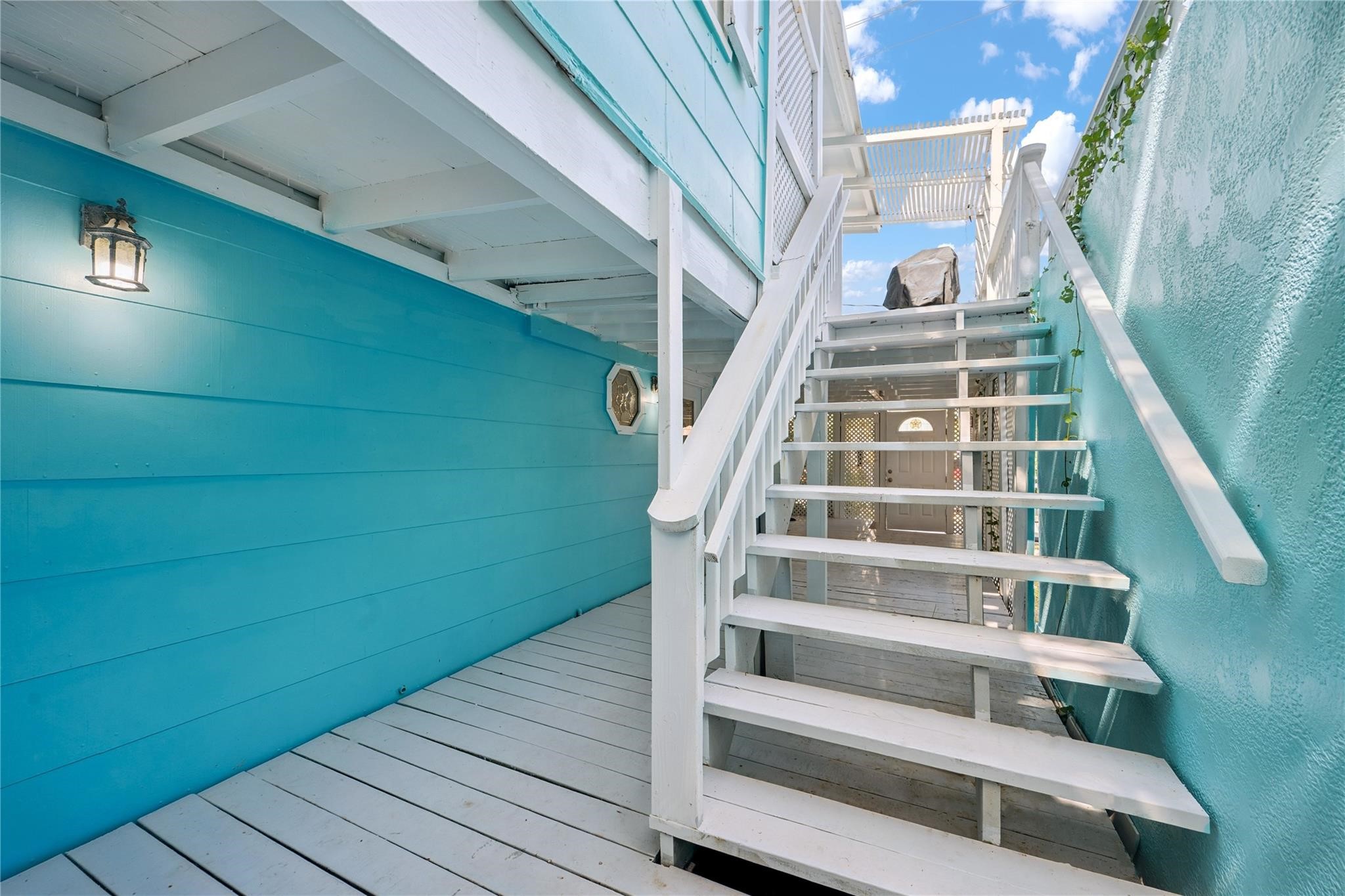 Exterior Staircase to Second Floor Upper Deck: This inviting exterior staircase, featuring crisp white wooden steps and railings, vibrant turquoise siding, and wall-mounted lighting provides convenient access to the second-floor deck, perfect for enjoying the relaxed beachside lifestyle.