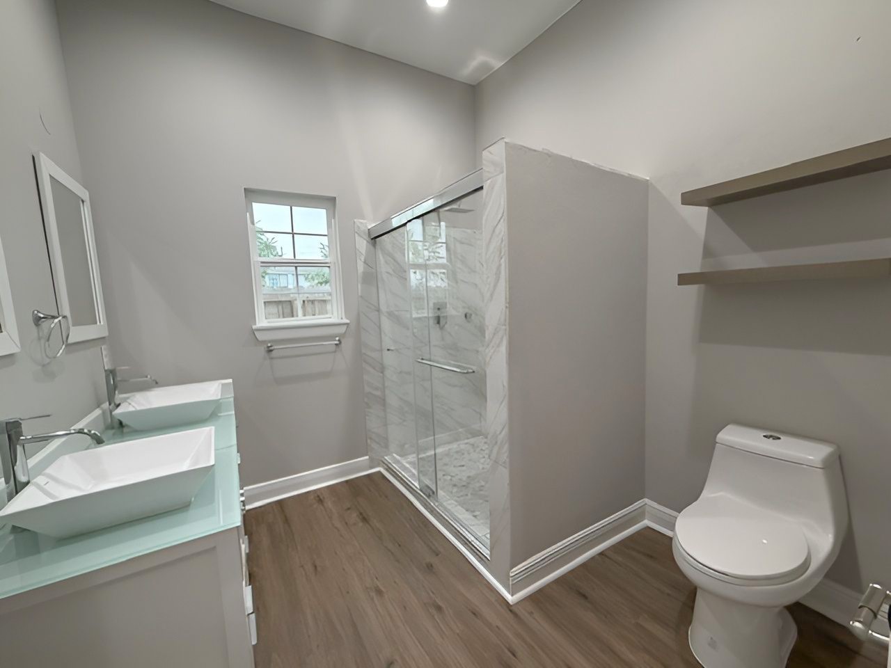 Spacious bathroom featuring a glass-enclosed walk-in shower with marble-look tile surround, modern fixtures, natural lighting, and convenient open shelving.