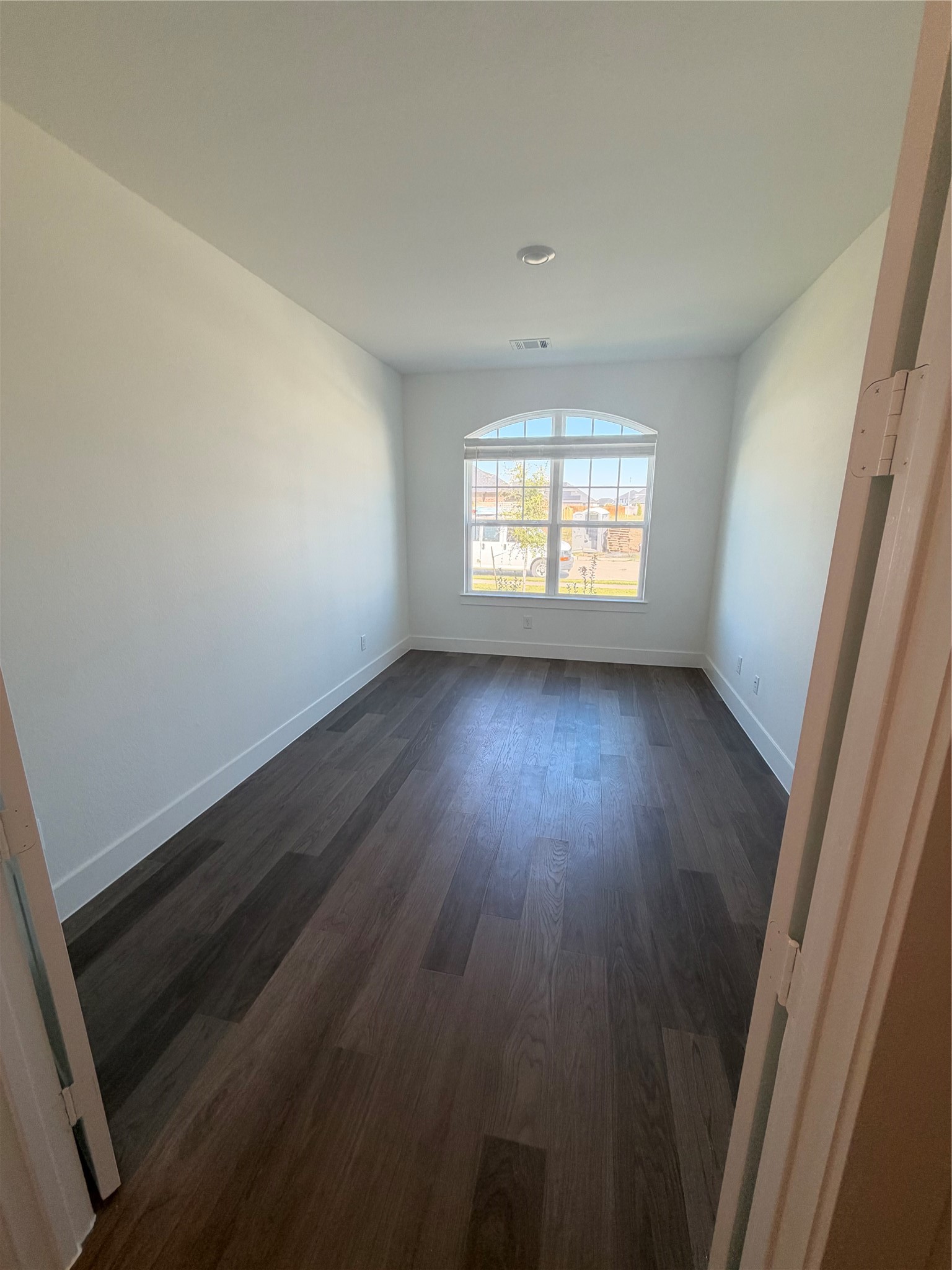 Bright office/den with elegant, dark wood flooring and a large arched window providing ample natural light.