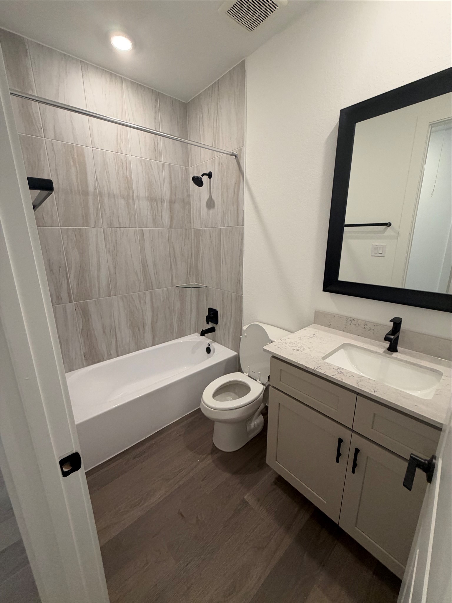 Modern bathroom featuring sleek wood flooring, a stylish vanity with a marble style countertop, and a tub-shower combo with elegant tile surround.