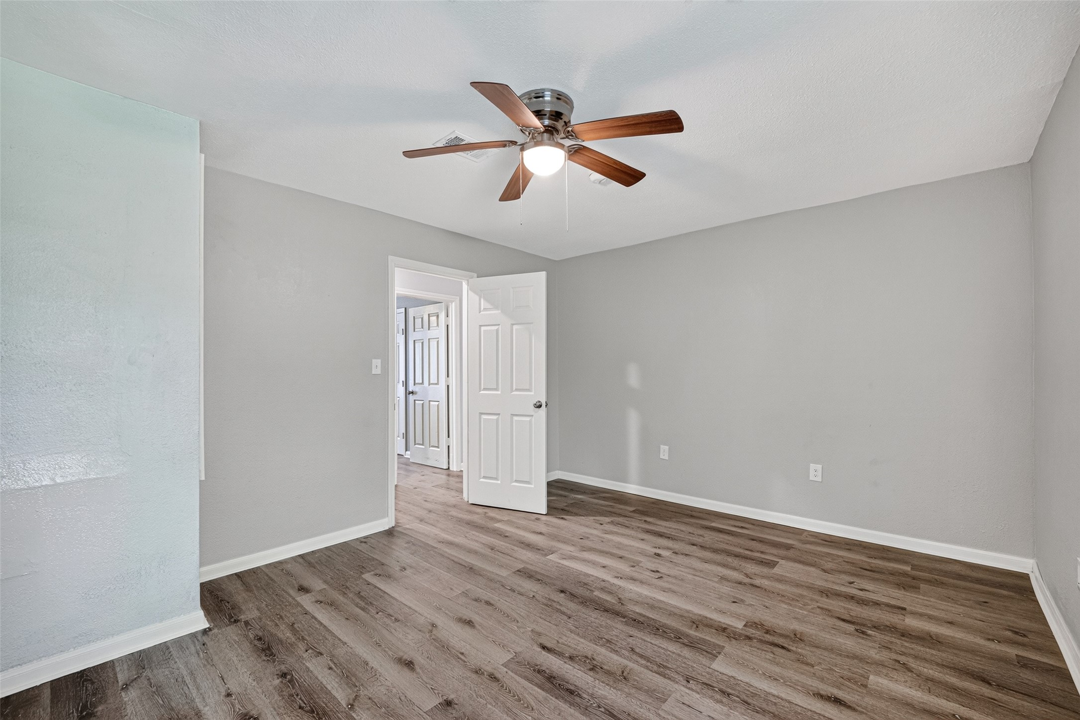 This room features neutral gray walls and wood-look flooring, complemented by a ceiling fan. The space is bright and open with a door leading to the hallway.